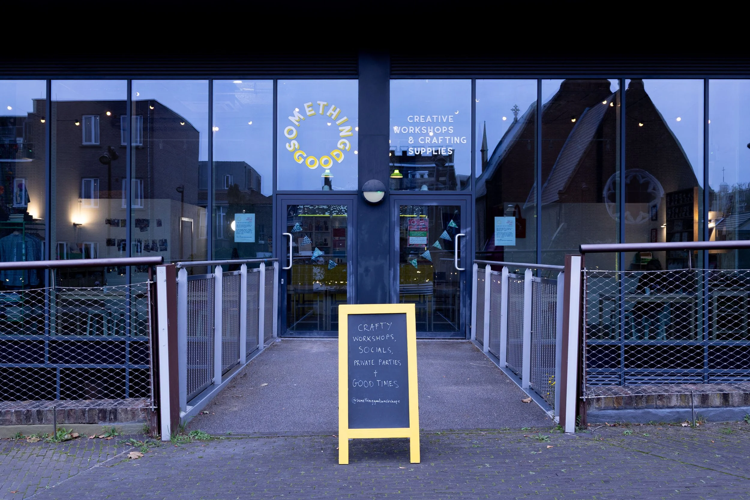 Storefront with large glass windows showing interior lights and reflection of buildings outside. Sign on door reads 'Something Good' and another sign outside lists offerings like craft workshops, socials, private parties, and good times.