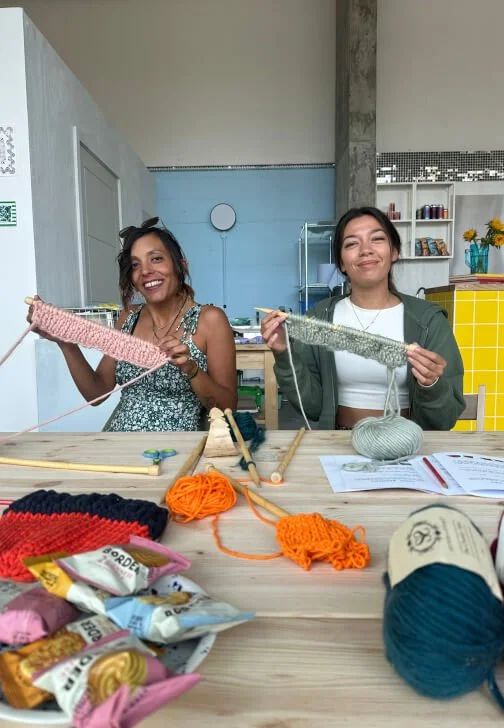 Two women sitting at a table, knitting with colorful yarn, surrounded by knitting supplies and snacks in a bright room.