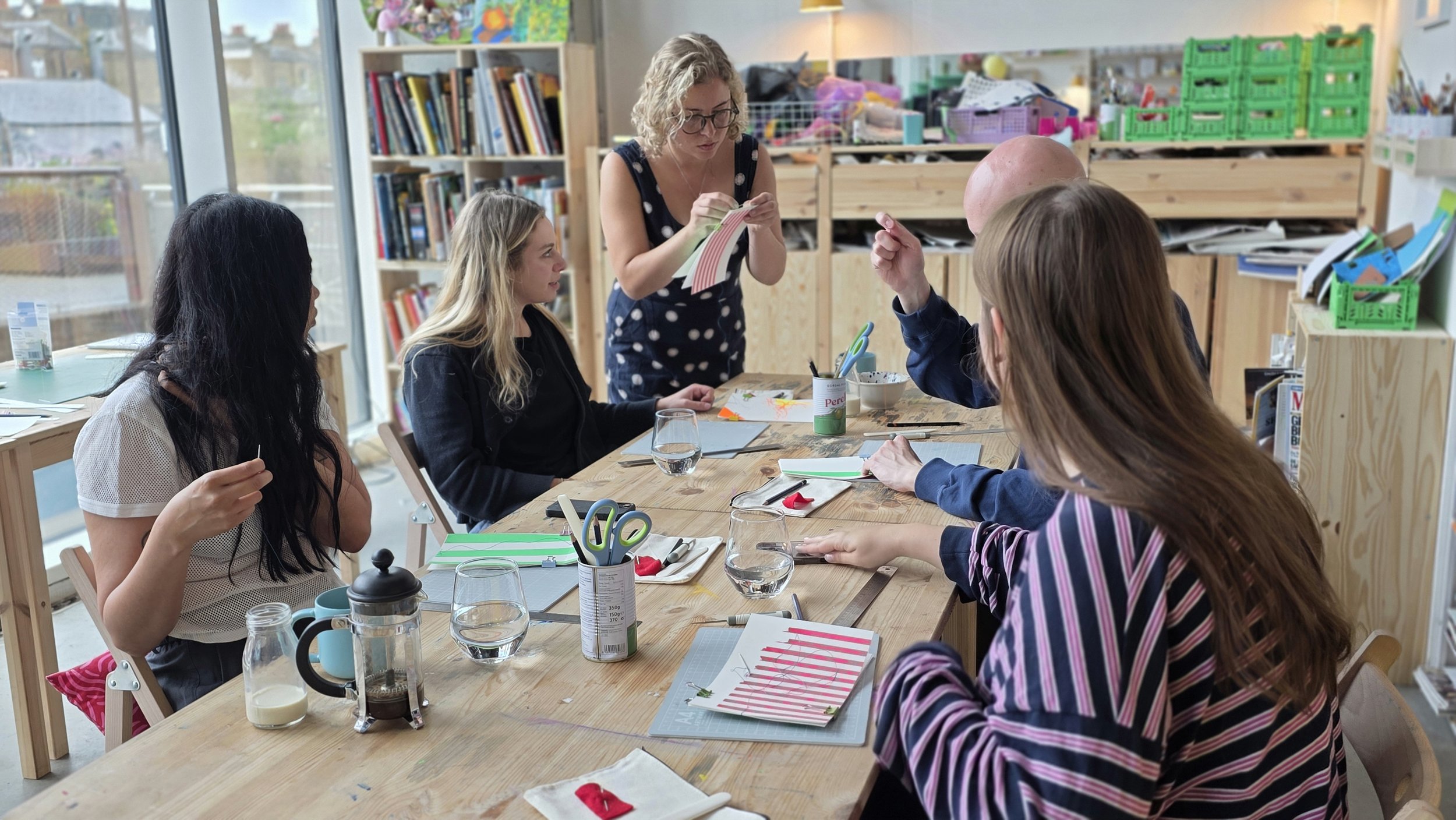 A group of five people sitting around a wooden table in a creative workspace, engaged in a bookbinding workshop, with bookshelves, colorful storage bins, and craft supplies in the background.