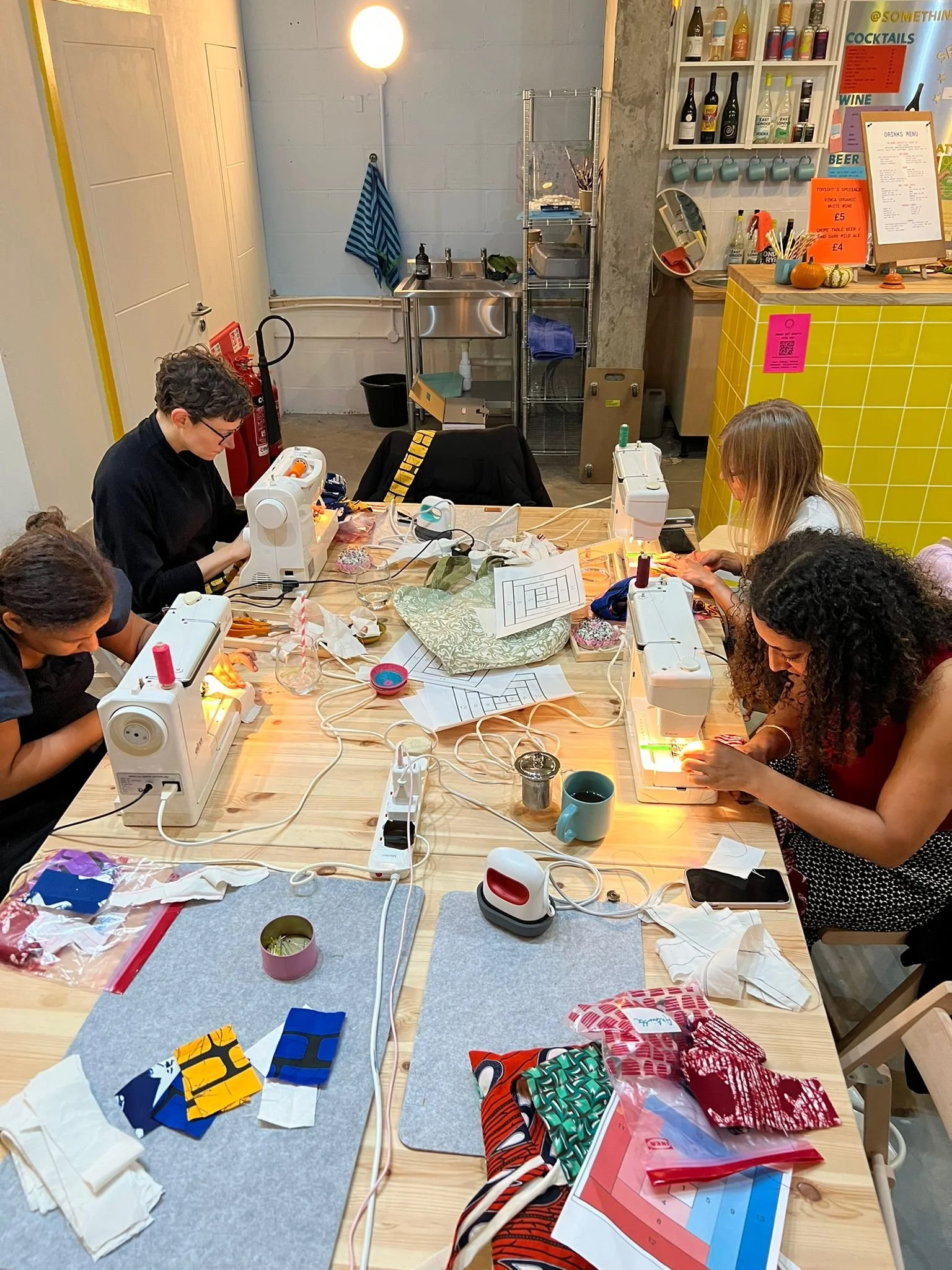 People working on sewing projects at a craft workshop in Deptford, London, with sewing machines and colorful fabric pieces on the table.