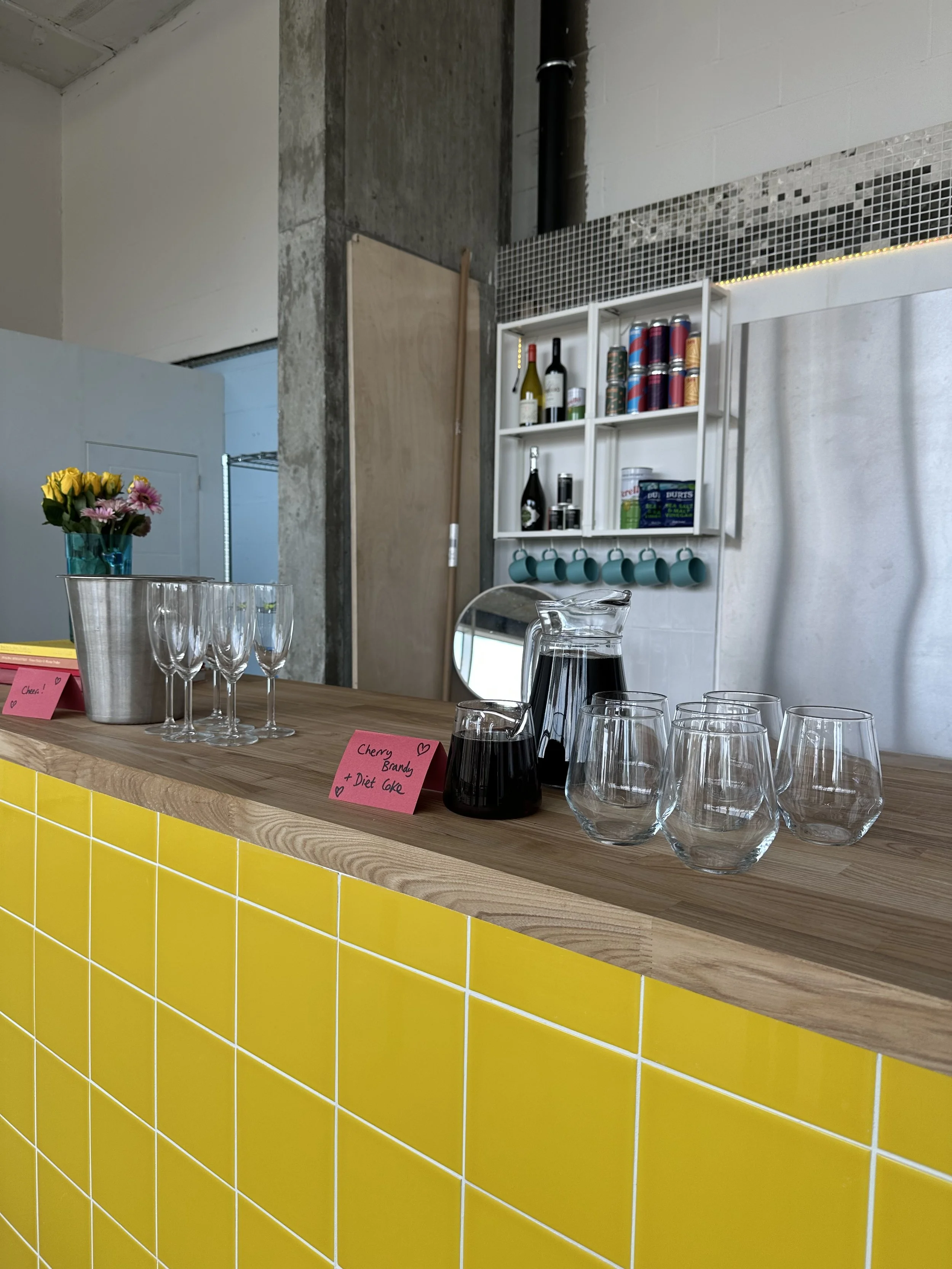 A bar counter with yellow tiled front, featuring four empty wine glasses, a small glass pitcher with dark red drink, a glass jug with dark liquid, a flowers in a small turquoise vase, and pink sticky notes, one labeled "Cherry Brandy + Diet Coke." In