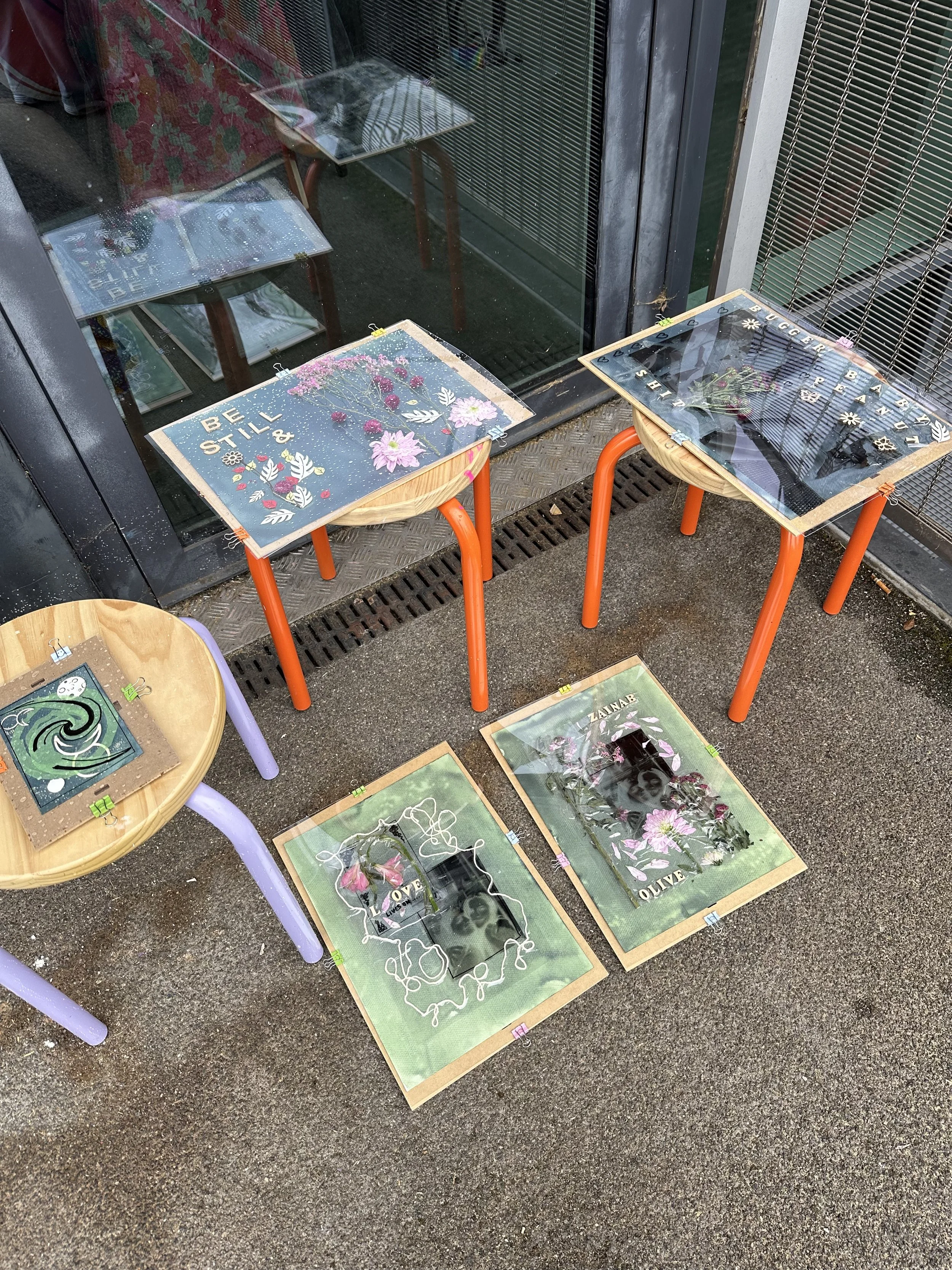 Outdoor display of art prints and posters on the ground and on small tables, with colorful floral and abstract designs, some with phrases like 'Be Still' and 'Olive'.