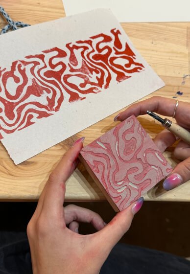 Person carving a marbled pattern into a small lino block with a carving tool at a craft workshop in London, with a paper print of the same pattern in front on a wooden table.