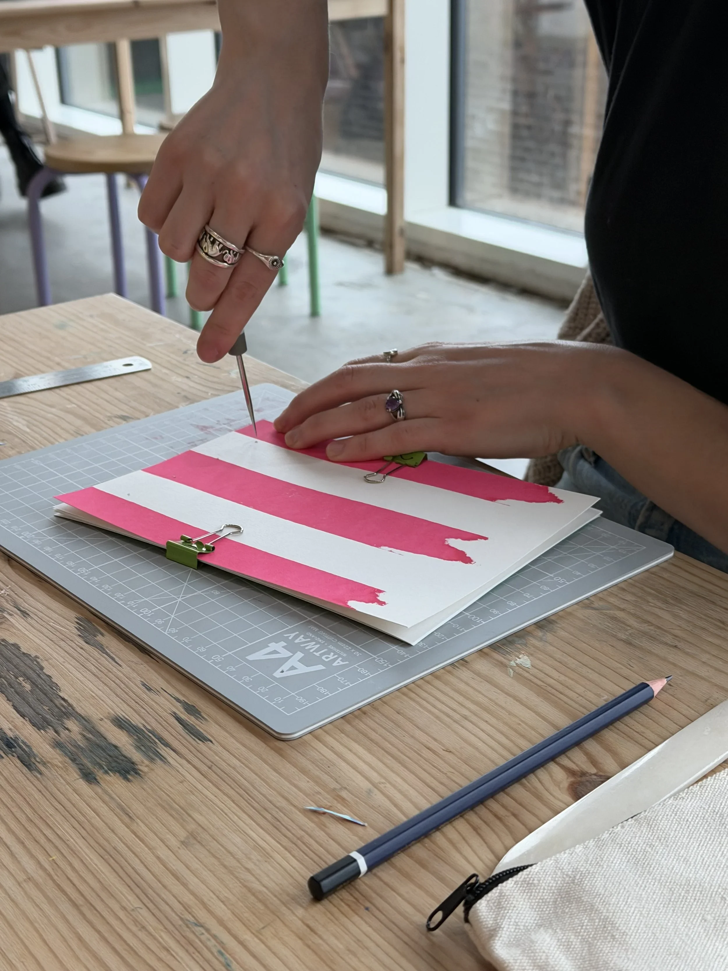 A person with multiple rings on their fingers is cutting paper with a craft knife on a workshop table. There are sheets with pink and white stripes, a metal ruler, a cutting mat, and a white bag nearby. The background shows large windows and a wooden structure.
