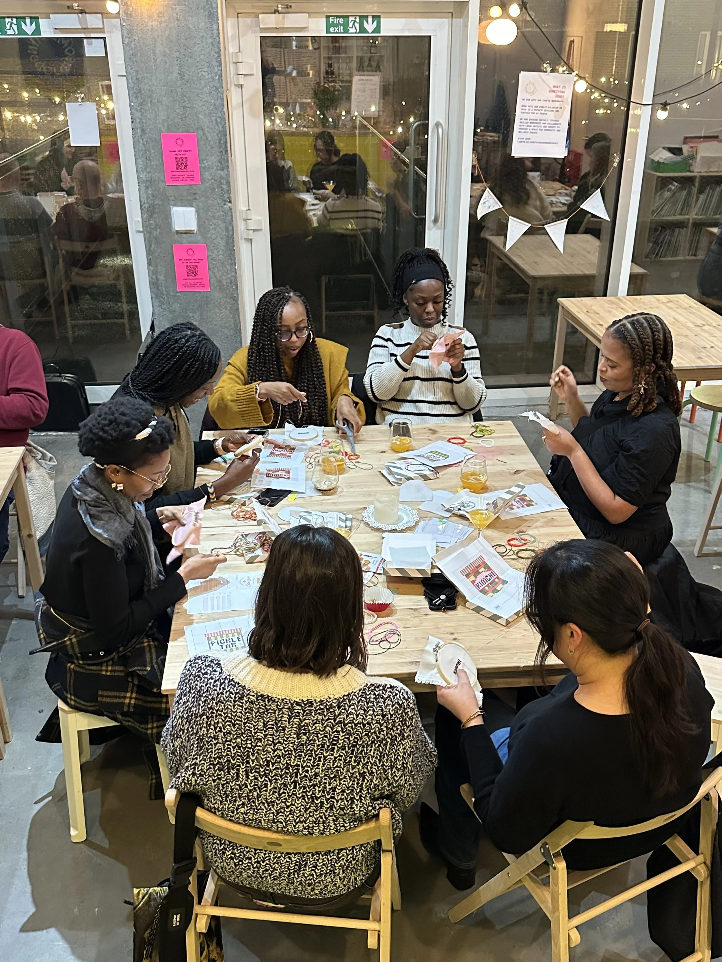 Two women holding fabric art pieces with botanical patterns, smiling inside a brightly lit room with large windows and a view of houses outside.