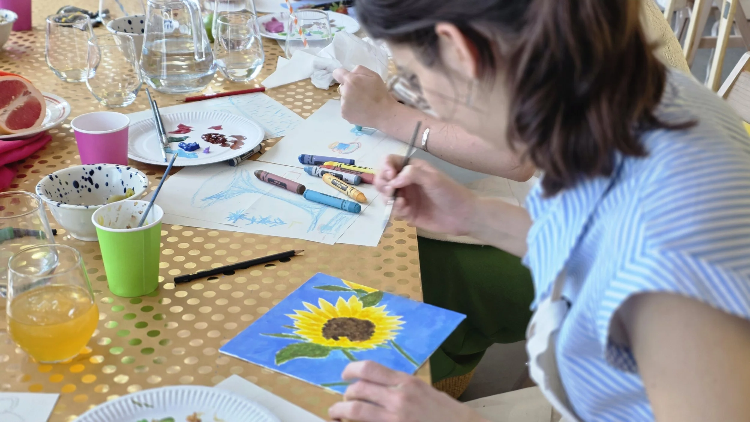 Person painting a sunflower on paper at a table with art supplies and drinks at a Hen Party at Something Good Workshops in Deptford.