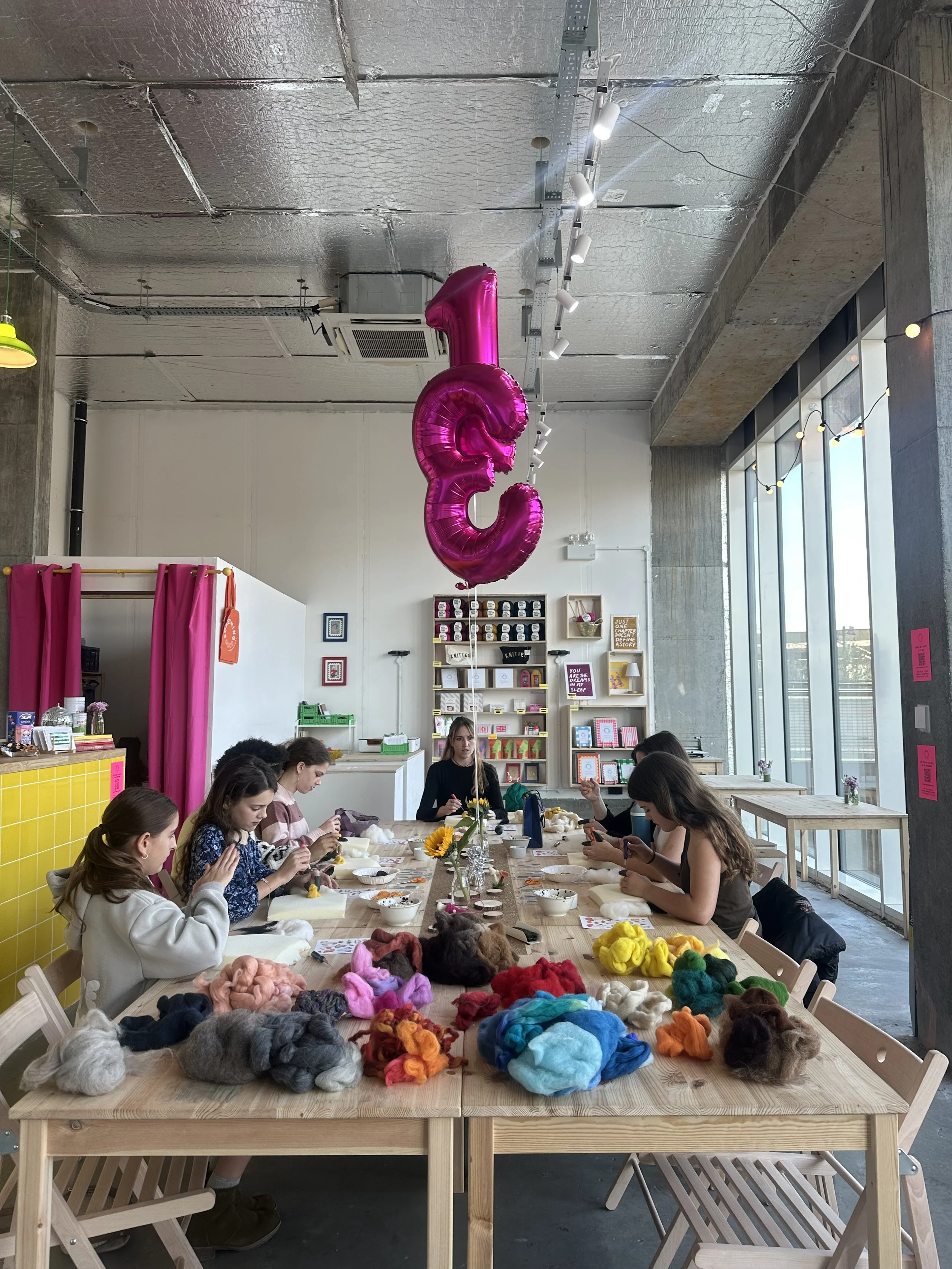 Several people, mostly children, sit at a large wooden table in a bright room with large windows. The table is covered with colorful wool or yarn, and some people are working on crafts or knitting. There are pink curtains on the left and shelves with various items, and a large pink balloon shaped like the numbers 1 and 3 hanging from the ceiling.