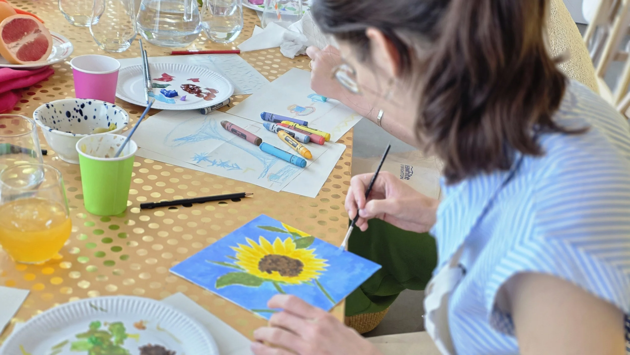 People engaging in a painting activity at a table with art supplies, including paints, brushes, and a painting of a sunflower at a Hen do painting class in London.