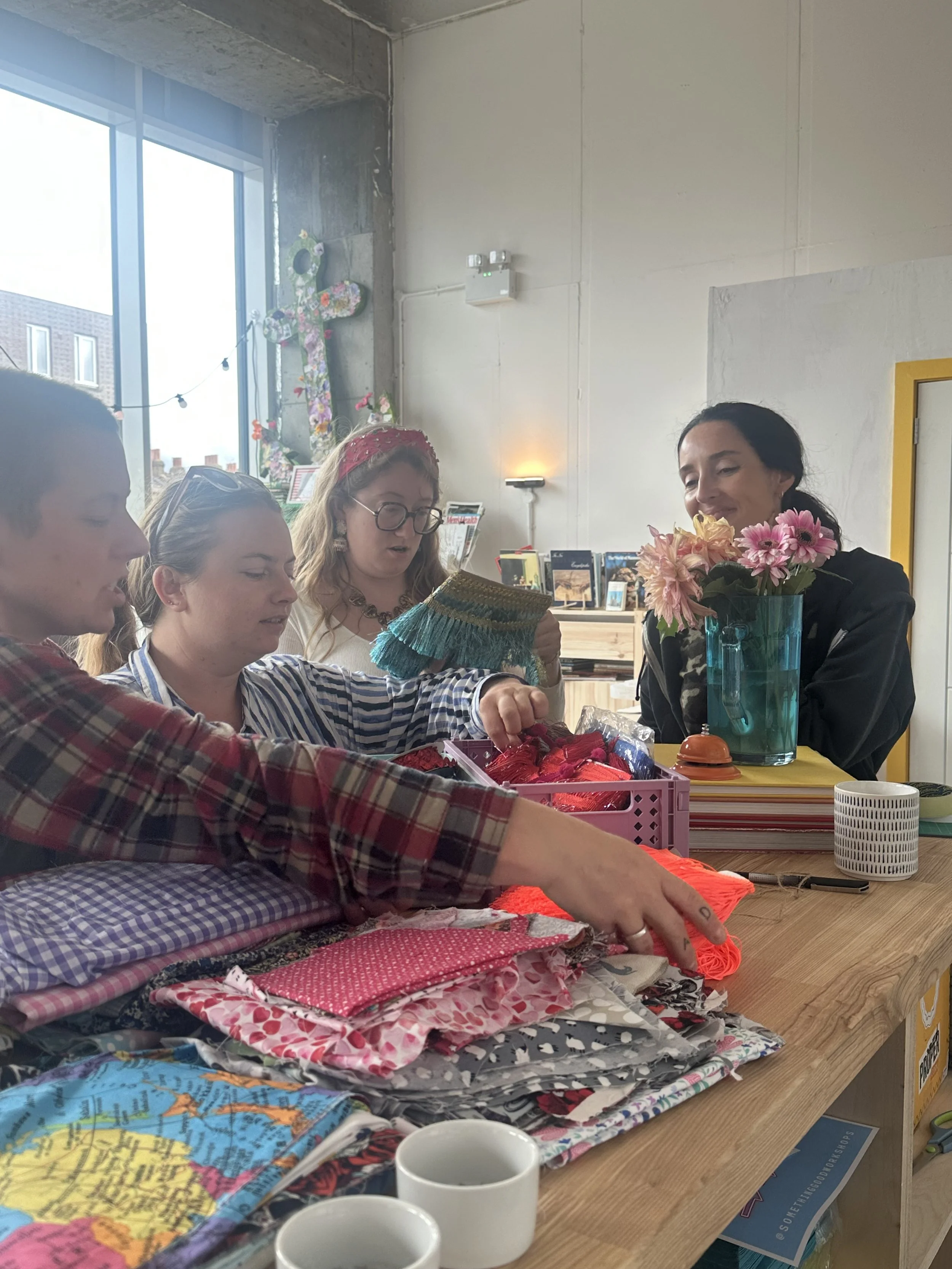 Group of women gathered around a table with colorful fabrics, yarn, and craft supplies, engaged in a craft activity at Something Good Workshops in Deptford, London, in a room with a large window, flowers, and books in the background.