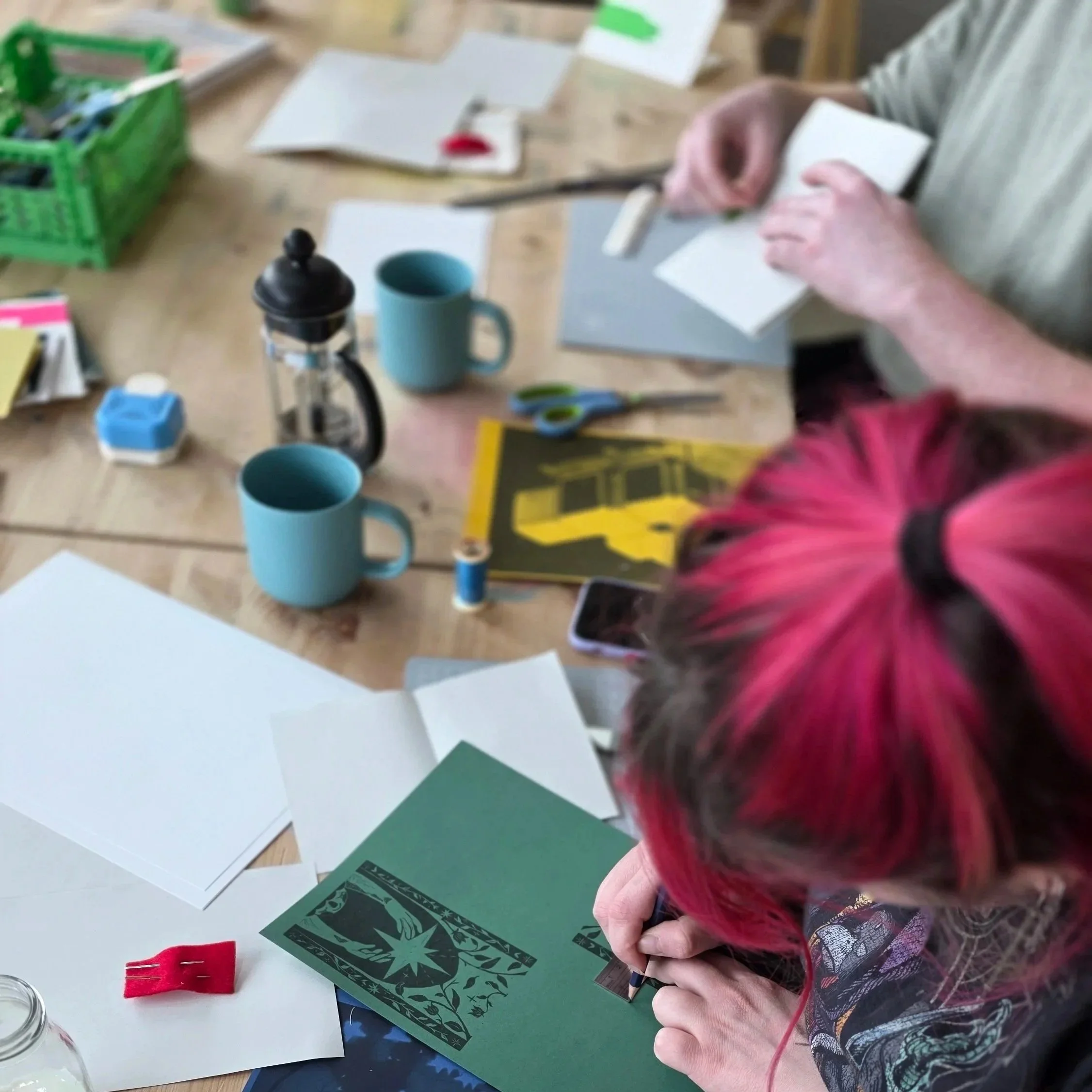 A person with long pink hair working at a table with various craft supplies, including paper, scissors, and a black card with a green design. There are also mugs, a small red felt piece, and other crafting tools visible on the table.