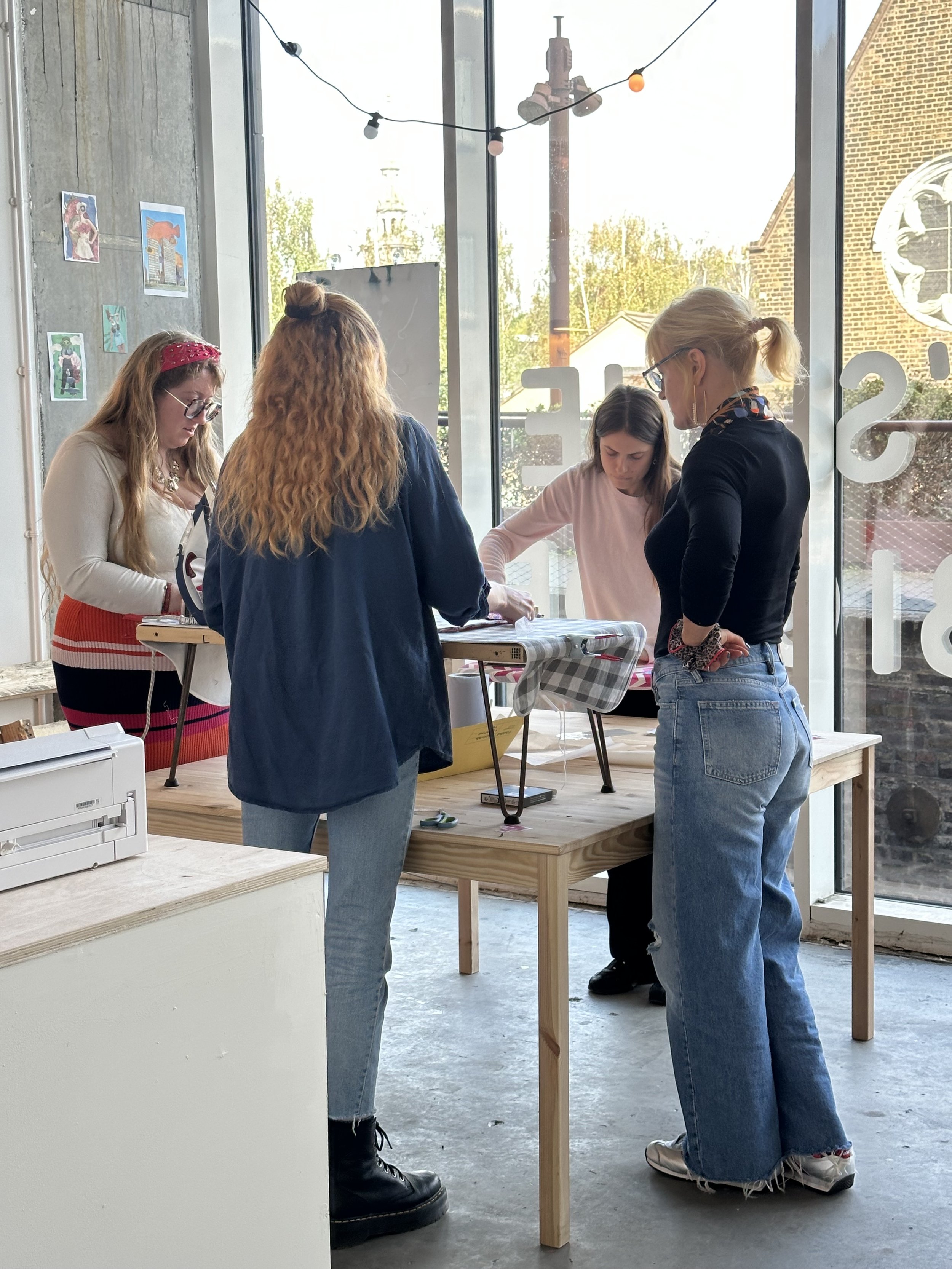 Four young women standing around a wooden table inside a bright modern space with large windows, engaged in a creative activity at Something Good Workshops in Deptford, London.