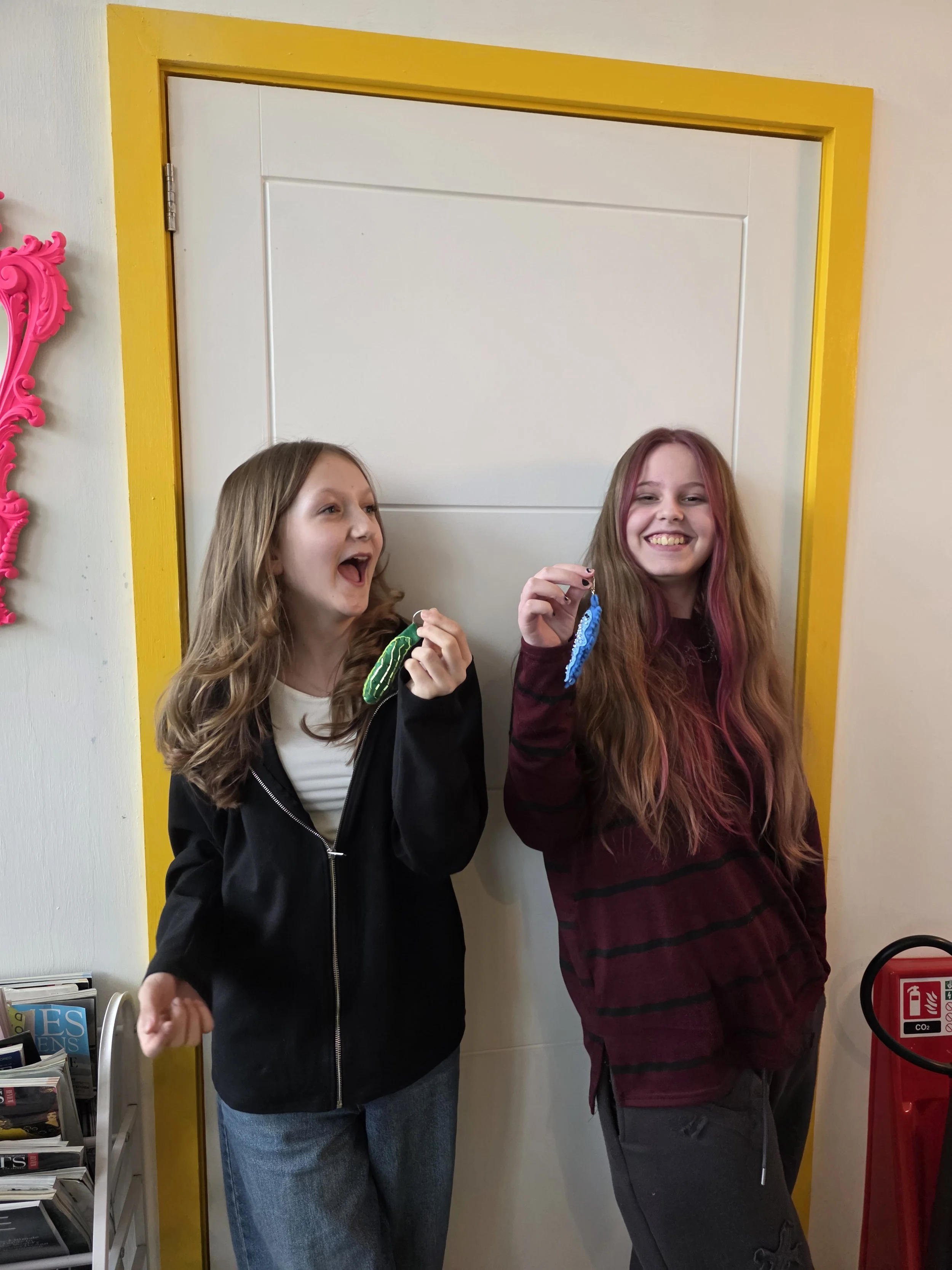 Two girls smiling and holding colorful felt charms, standing in front of a white door with yellow trim.