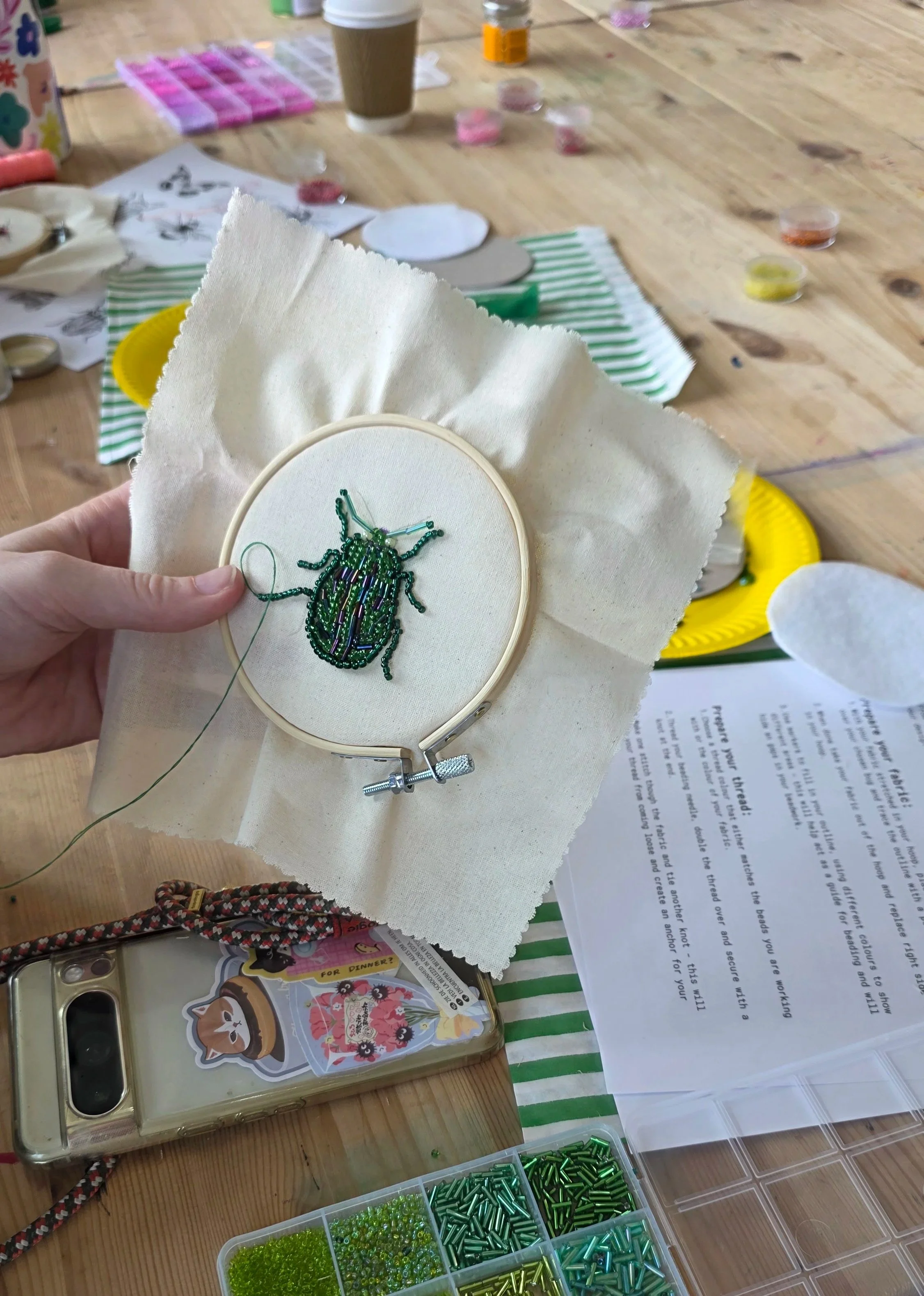A hand holding a sewing hoop with a partially completed beaded beetle embroidery. The work is on white fabric, surrounded by a variety of tools and materials including containers of beads in different shades of green, pink, and yellow, a coffee cup, printed instructions, and a colorful phone case on a wooden table.
