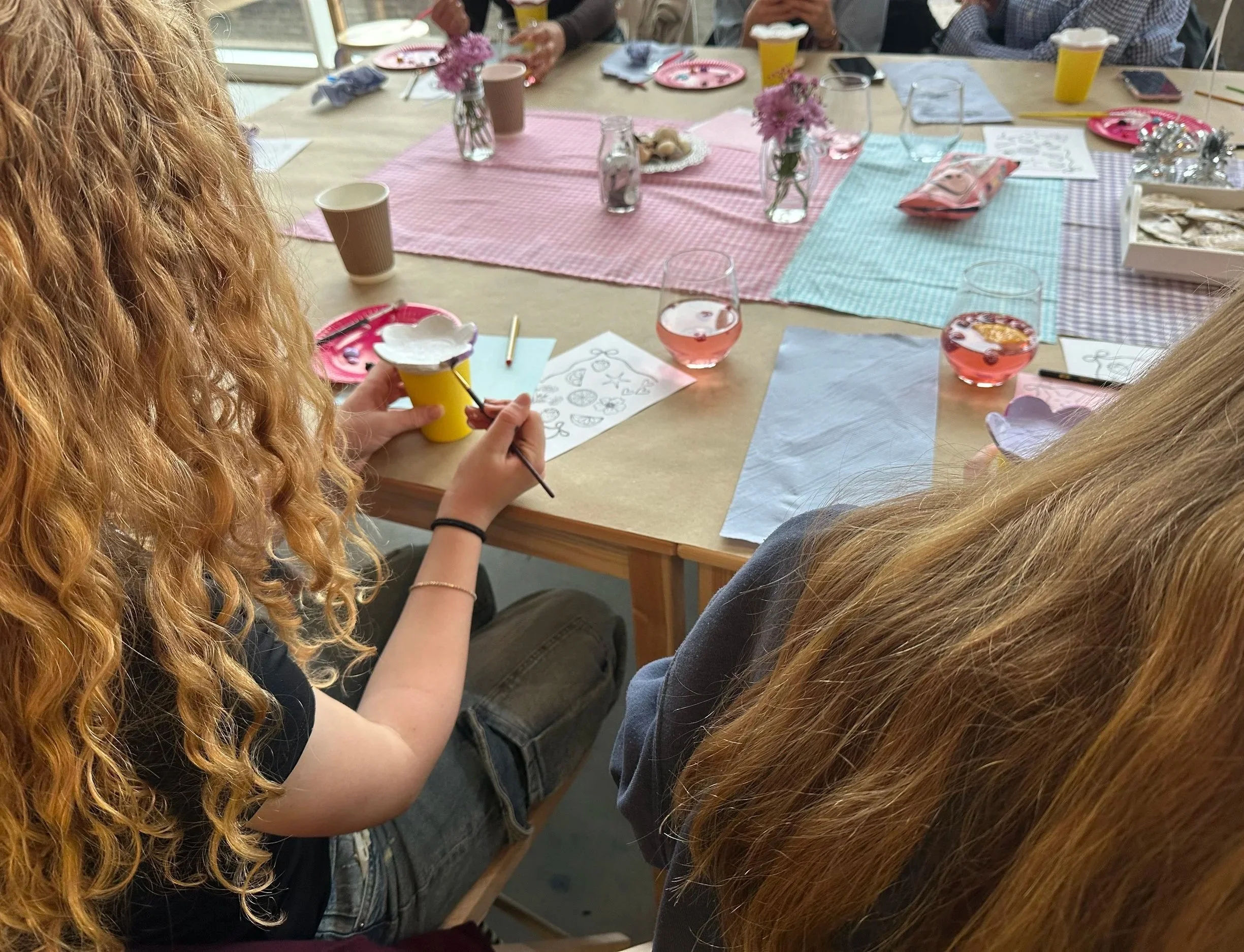 People sitting around a table decorated with pink and blue checkered tablecloths, pink flowers in glass vases, and pink cups, enjoying a social gathering or celebration.
