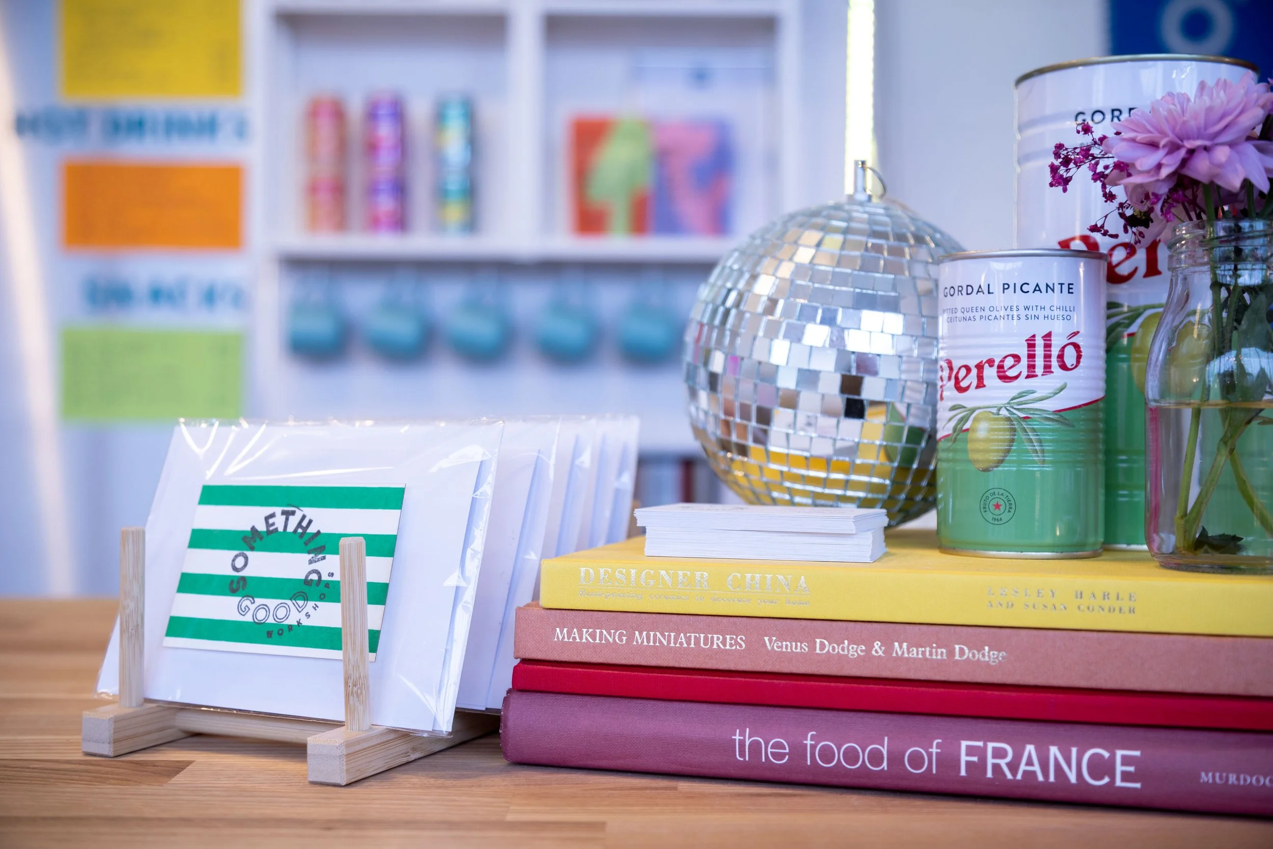 Close-up of books, a decorated disco ball, cans with flowers, and a sign on a wooden table in a display or store.