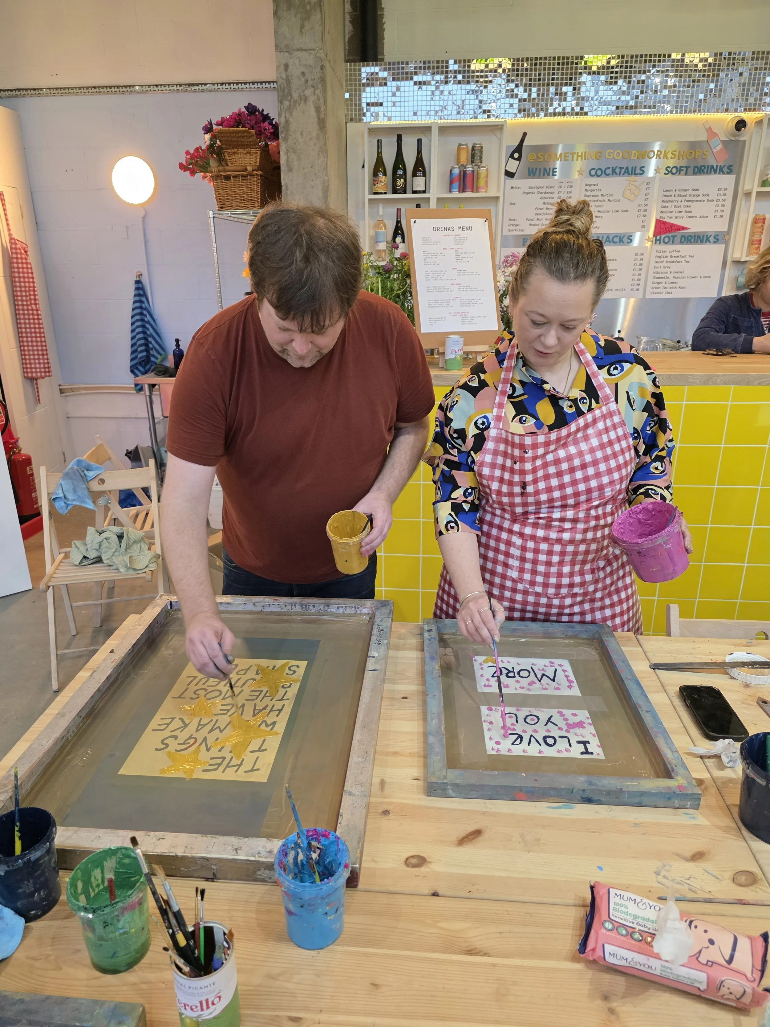 Two people working on screen printing art projects at a workshop, with various paint brushes and supplies on the wooden table.