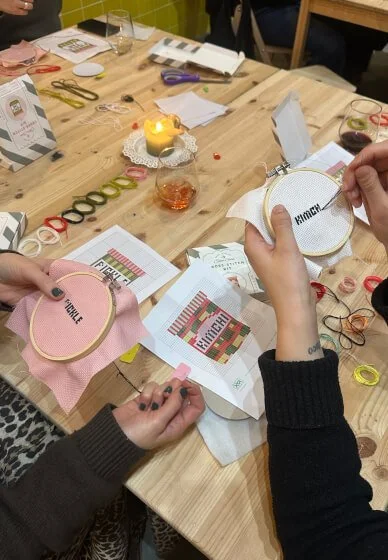 People crafting embroidery projects at a wooden table with embroidery hoops, colorful thread, scissors, and sewing patterns.