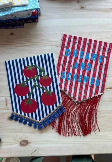 Two fabric pennants with decorative fringes, one with a tomato design on blue and white stripes, and the other with the words 'DREAMS AND SCHEMES' on red and white stripes, placed on a light wooden surface.