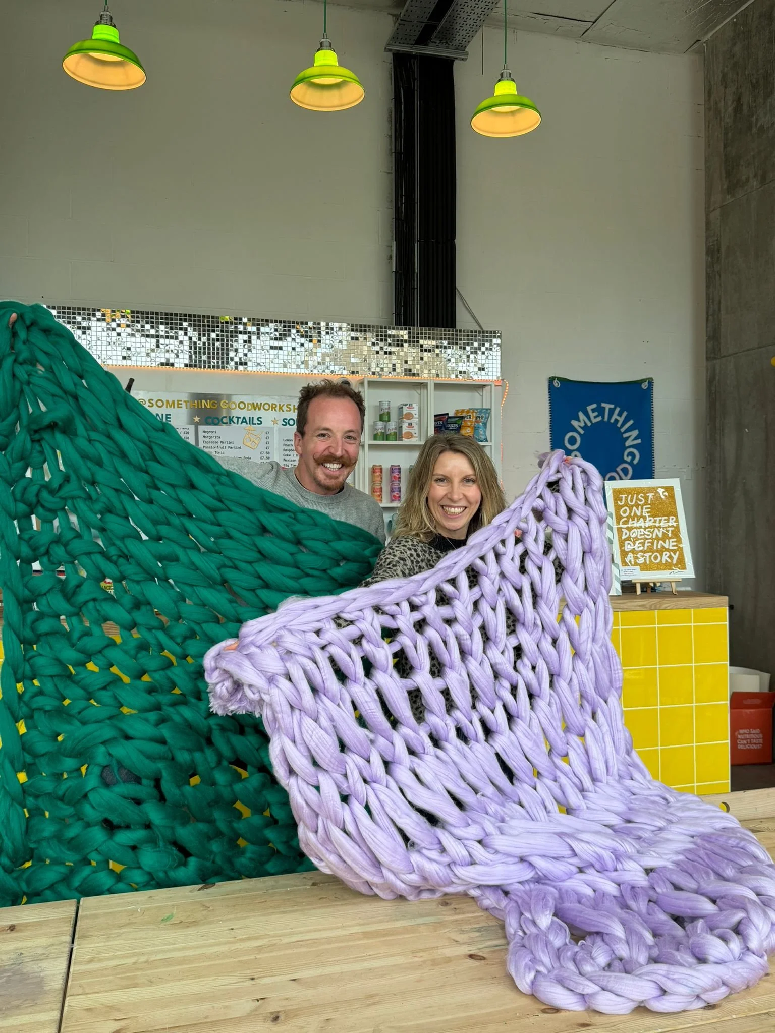 Two smiling people posing behind large, colorful knitted or woven furniture resembling oversized chairs or loungers in green and purple, inside a modern space with industrial ceiling lighting and a sign that reads 'Nothing Goodworkshs' in the backgro