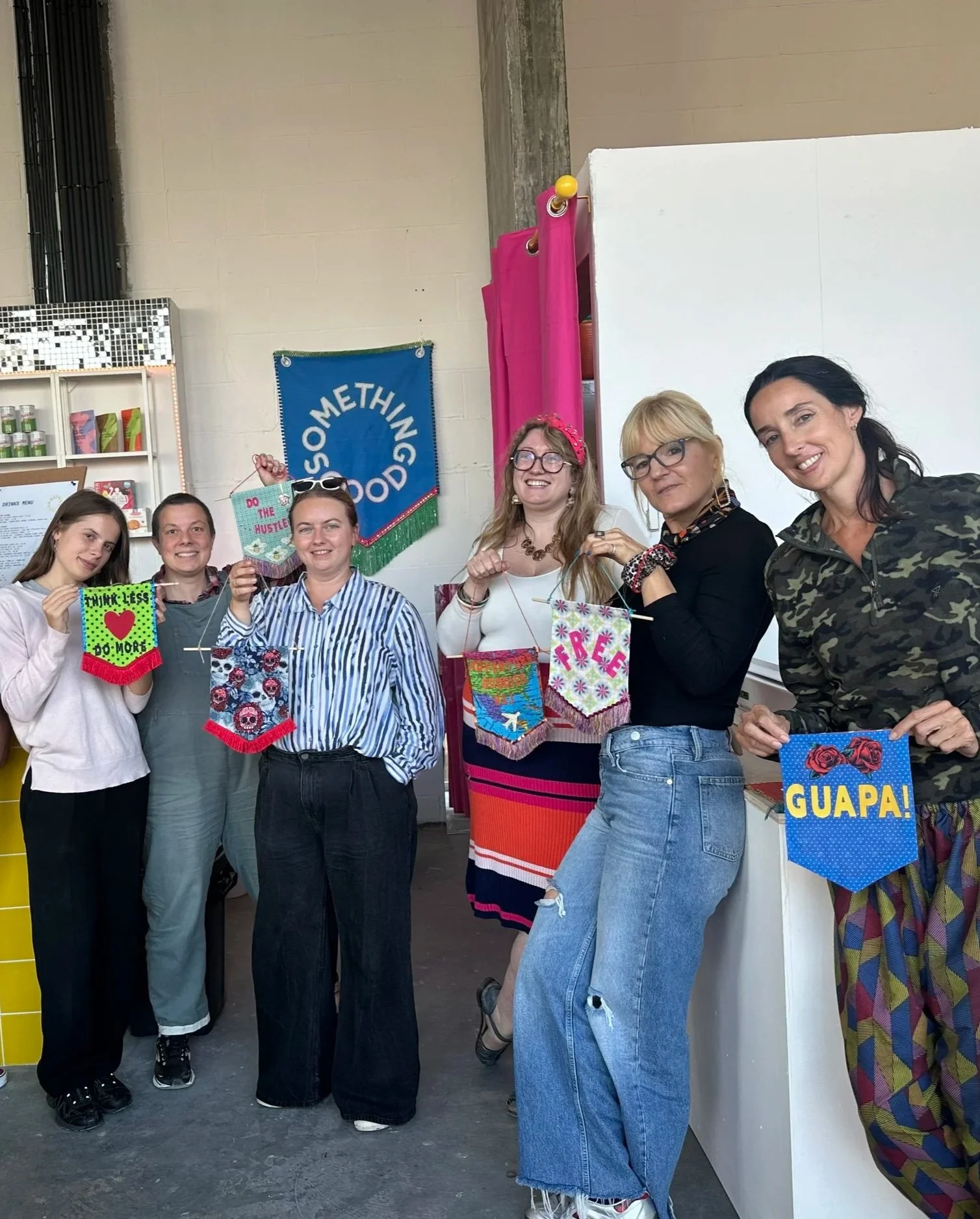 Group of six women holding colorful small banners with positive messages, smiling, at a craft workshop with a blue banner that says 'SOMETHING GOOD', a pink fabric backdrop, and a white wall.