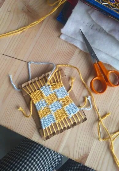 Small weaving loom with yellow and white checkered pattern, yellow yarn, and scissors on a wooden table.
