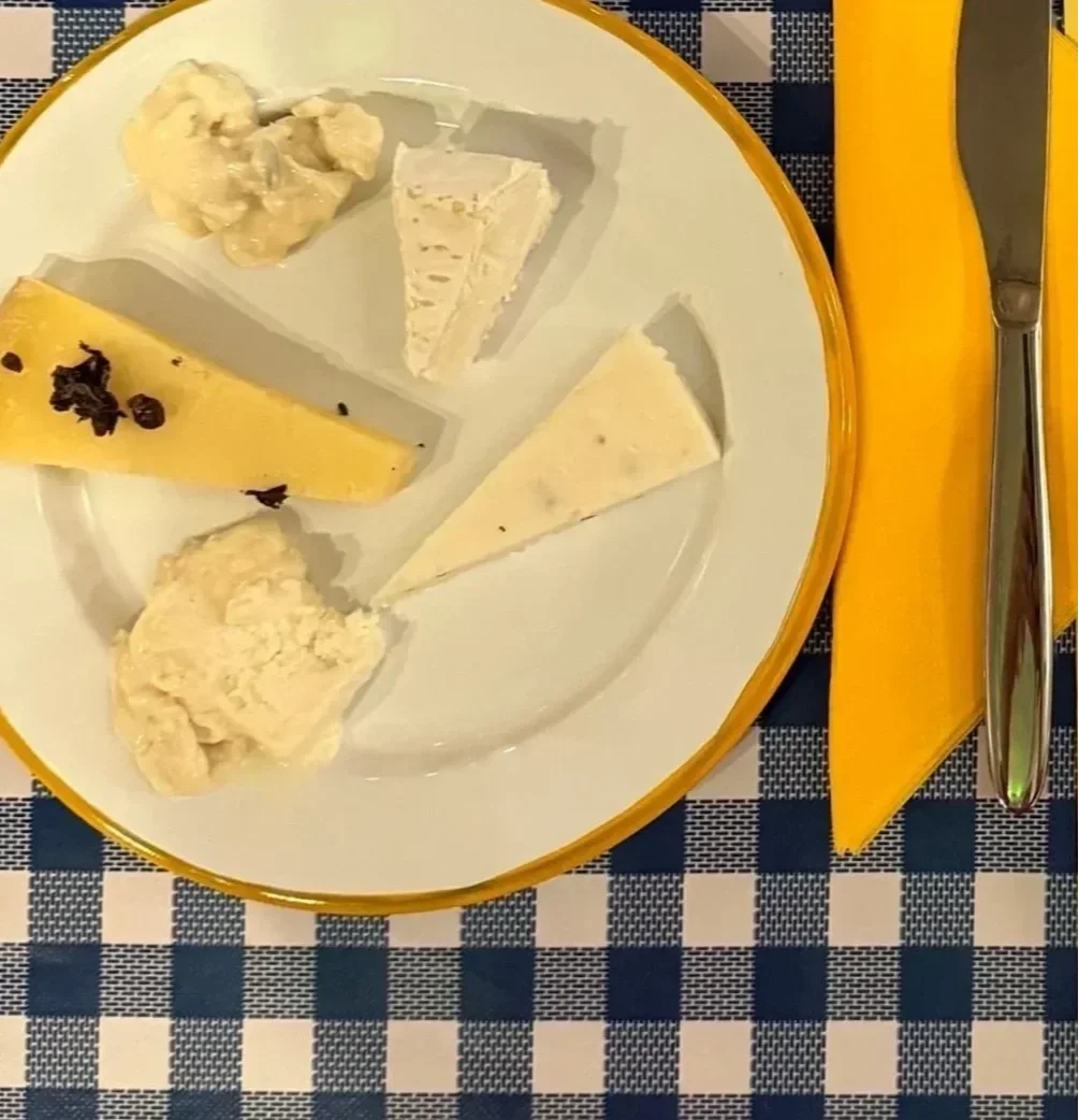 A plate with five different types of cheese arranged around the edge, on a blue and white checkered tablecloth, with a yellow napkin, knife, and spoon beside it.