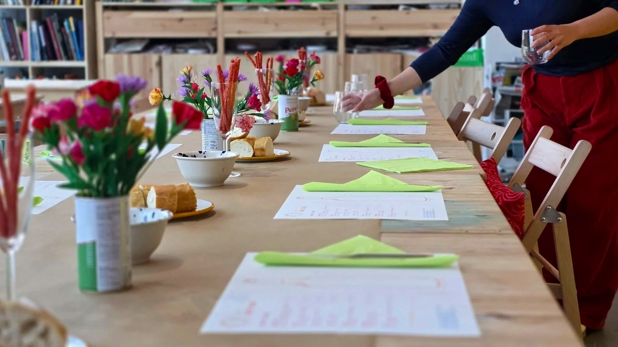 A long wooden table set with colorful flower arrangements, plates of bread, green cloth napkins, glasses, and printed menus, in a cozy room with wooden shelves in the background. A person is reaching across the table.