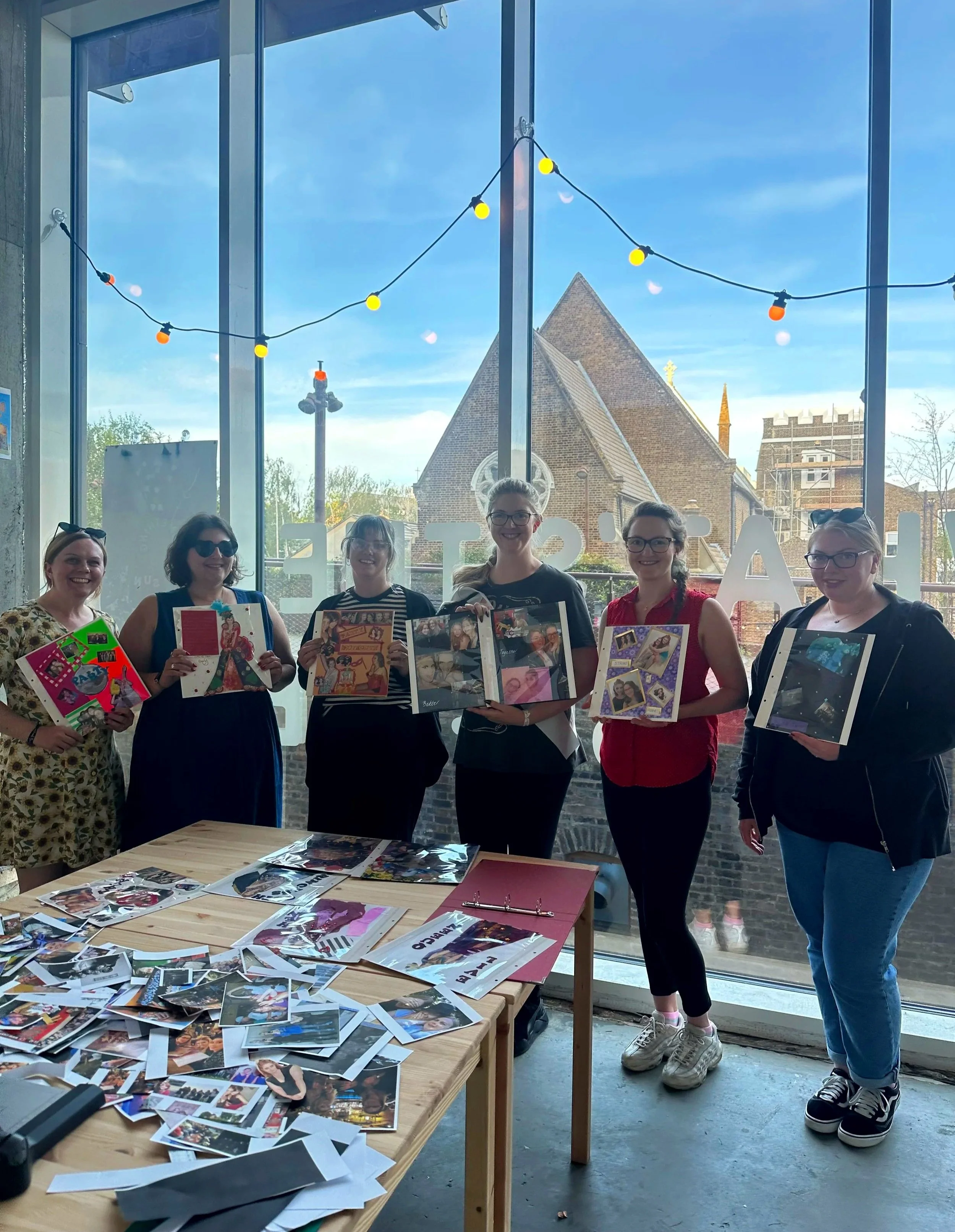 Five women standing indoors in front of a large window holding up colorful scrapbook pages or photo albums, with a table in front covered in scattered photographs and papers.