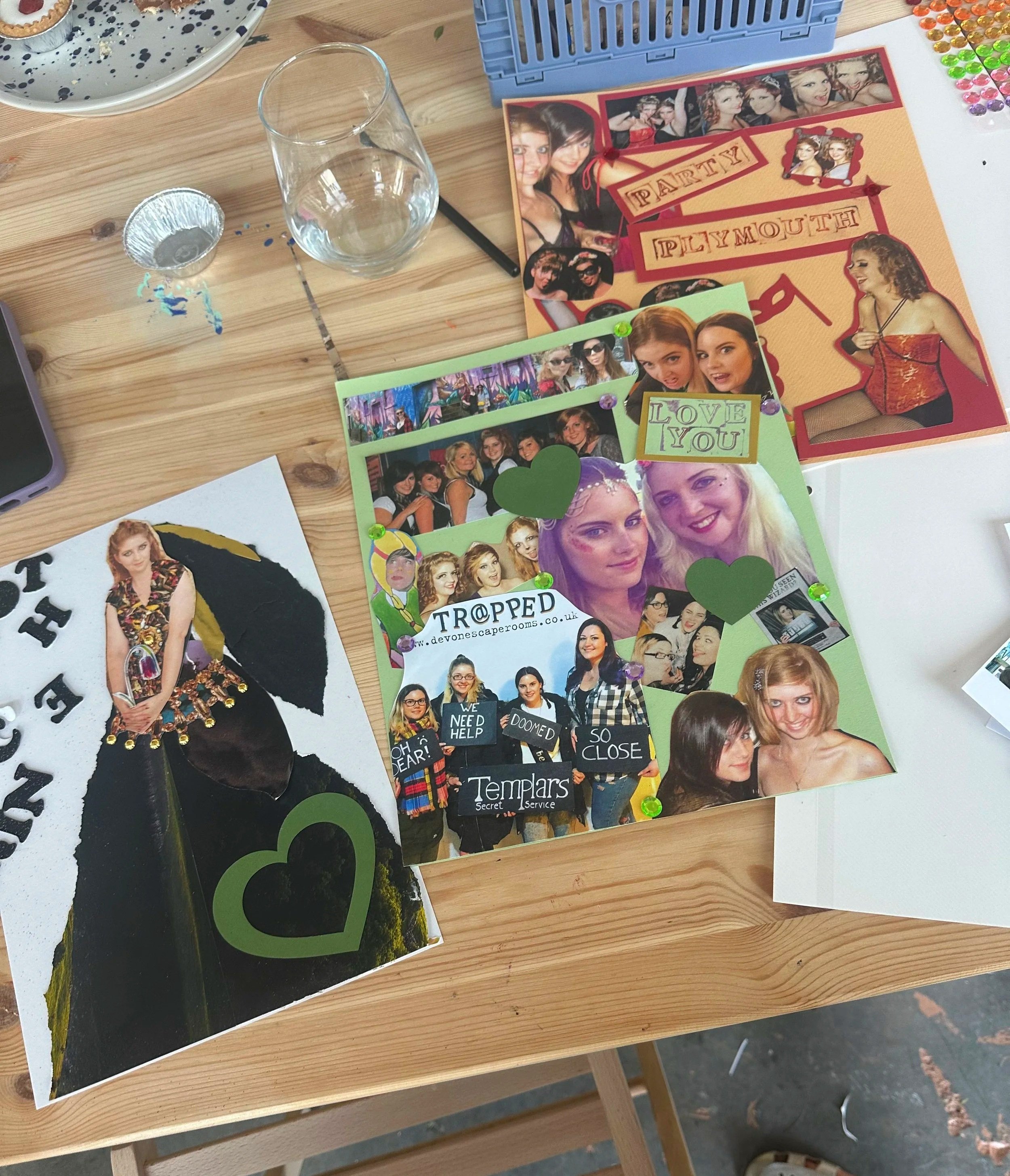 Photo of a wooden table with various photos and craft supplies, including a blue container, colorful stickers, a glass, a cupcake tin, and photographs of women at a party and friends.
