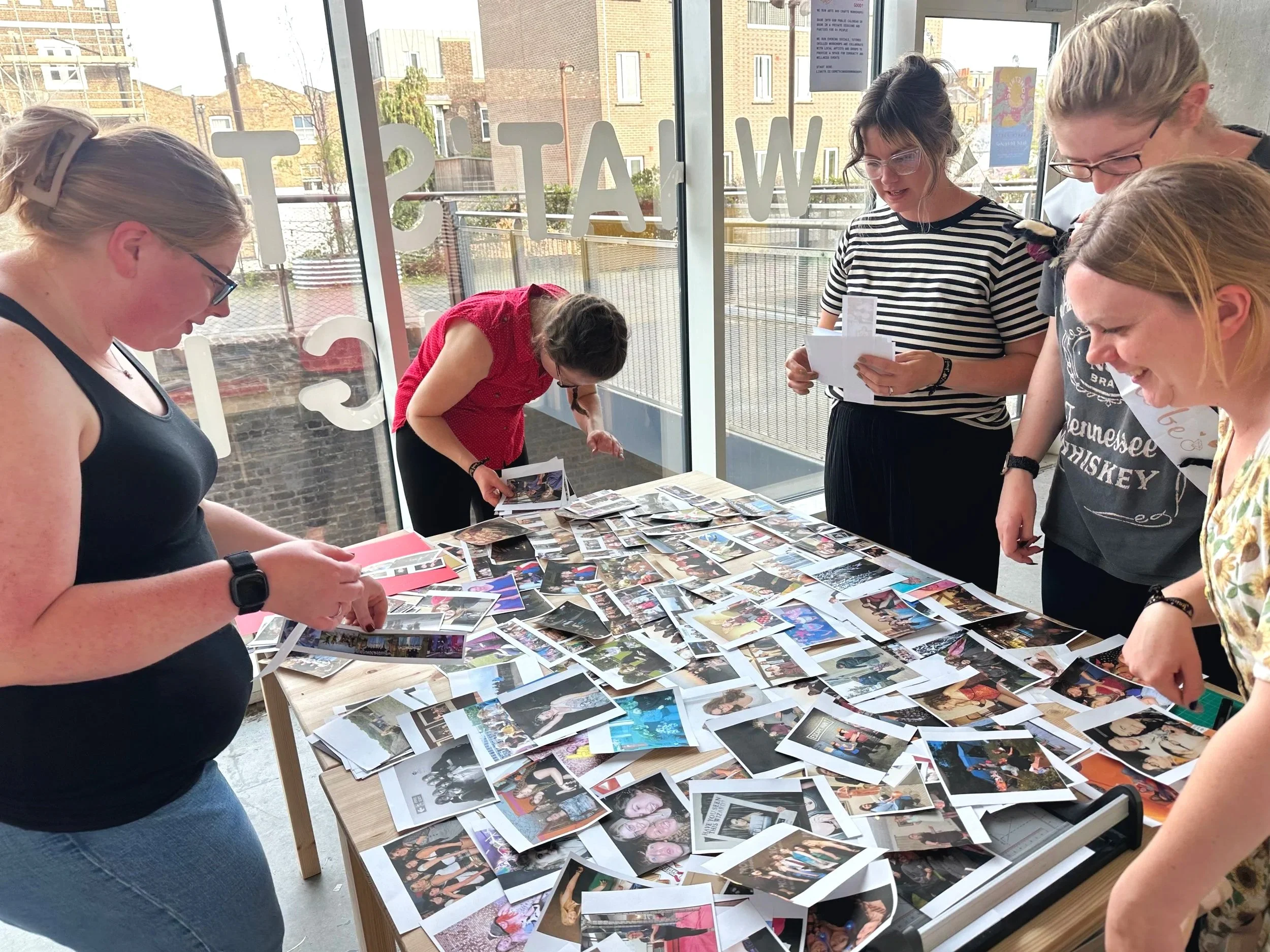 A group of people looking at numerous printed photographs spread out on a table at a craft workshop space with large glass windows.