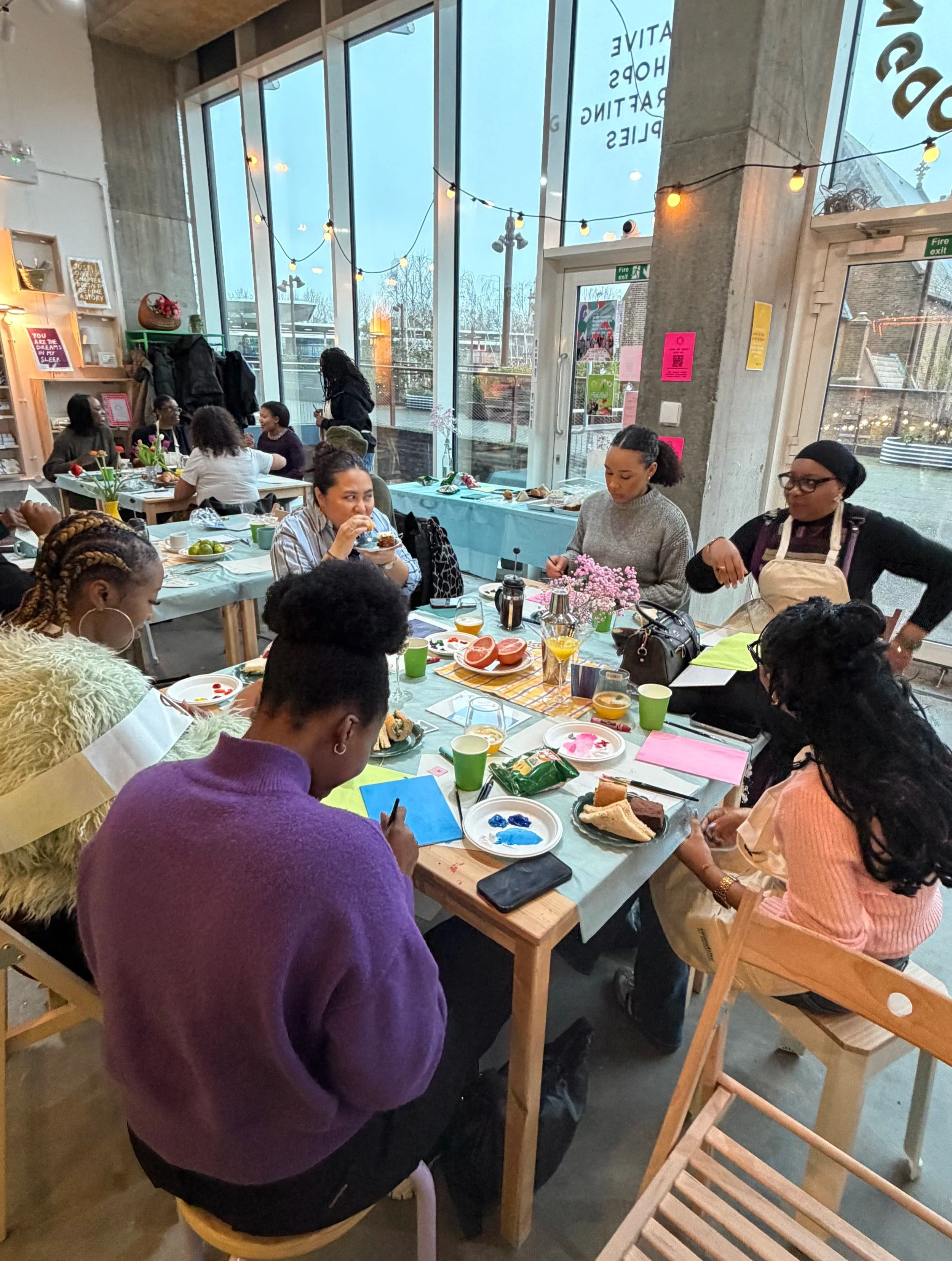A group of women gathered around a long table inside a modern craft workshop, eating and talking. The cafe has large windows, string lights, and a mix of wooden and concrete decor. The table has various plates of food, drinks, and decorative flowers.