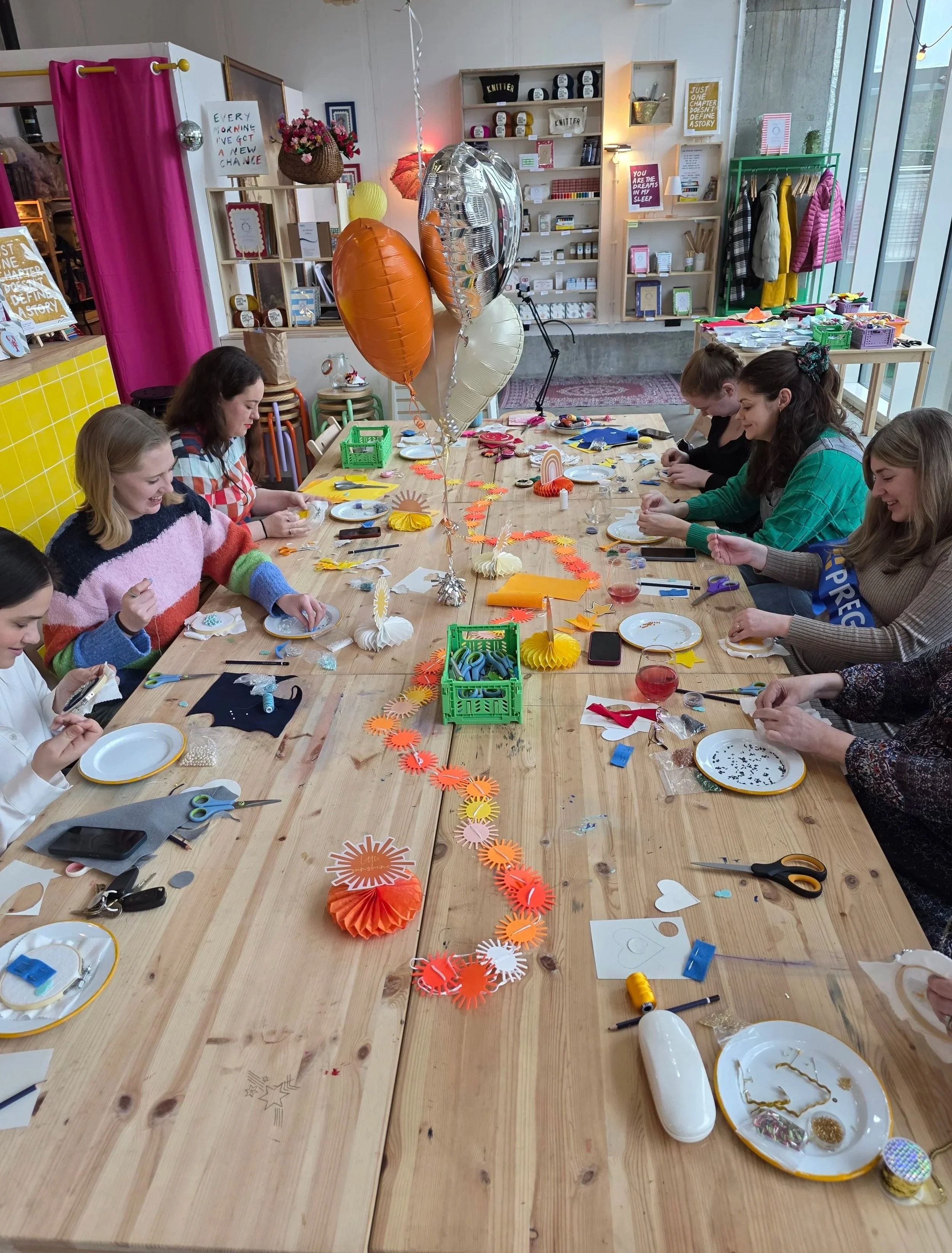 People sitting around a wooden table making paper felt charms at a craft workshop in London with balloons and decorations celebrating a baby shower in a craft room.