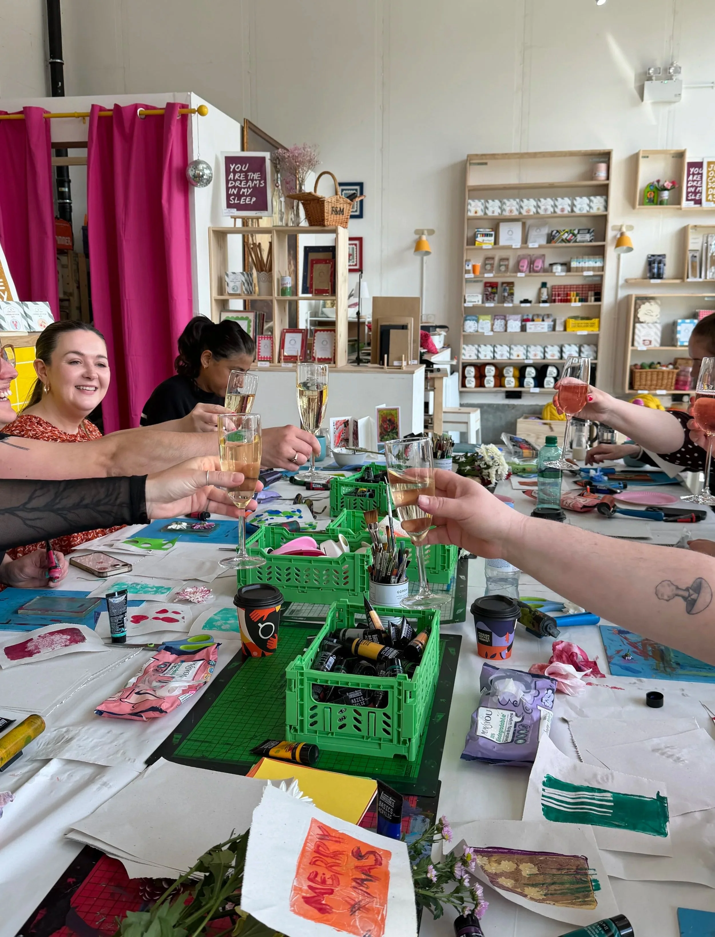 A group of people celebrating with drinks at a craft or art workshop, with art supplies, flowers, and colorful decorations on the table and shelves in the background.