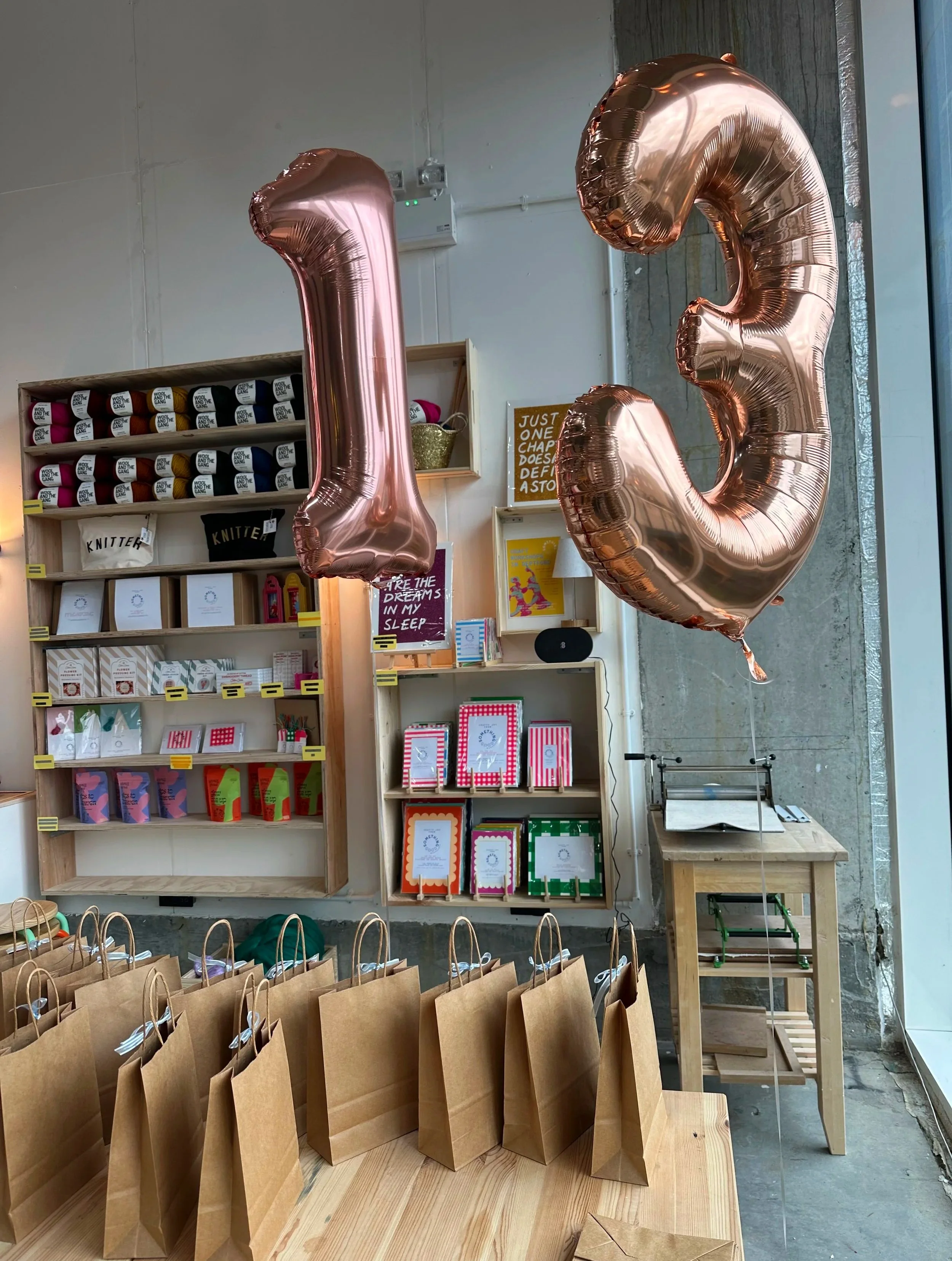 Brown paper grocery bags on a wooden table, with balloons shaped as the number three in rose gold foil decoration in the background at a teen birthday party at Something Good Workshops, Deptford, London.