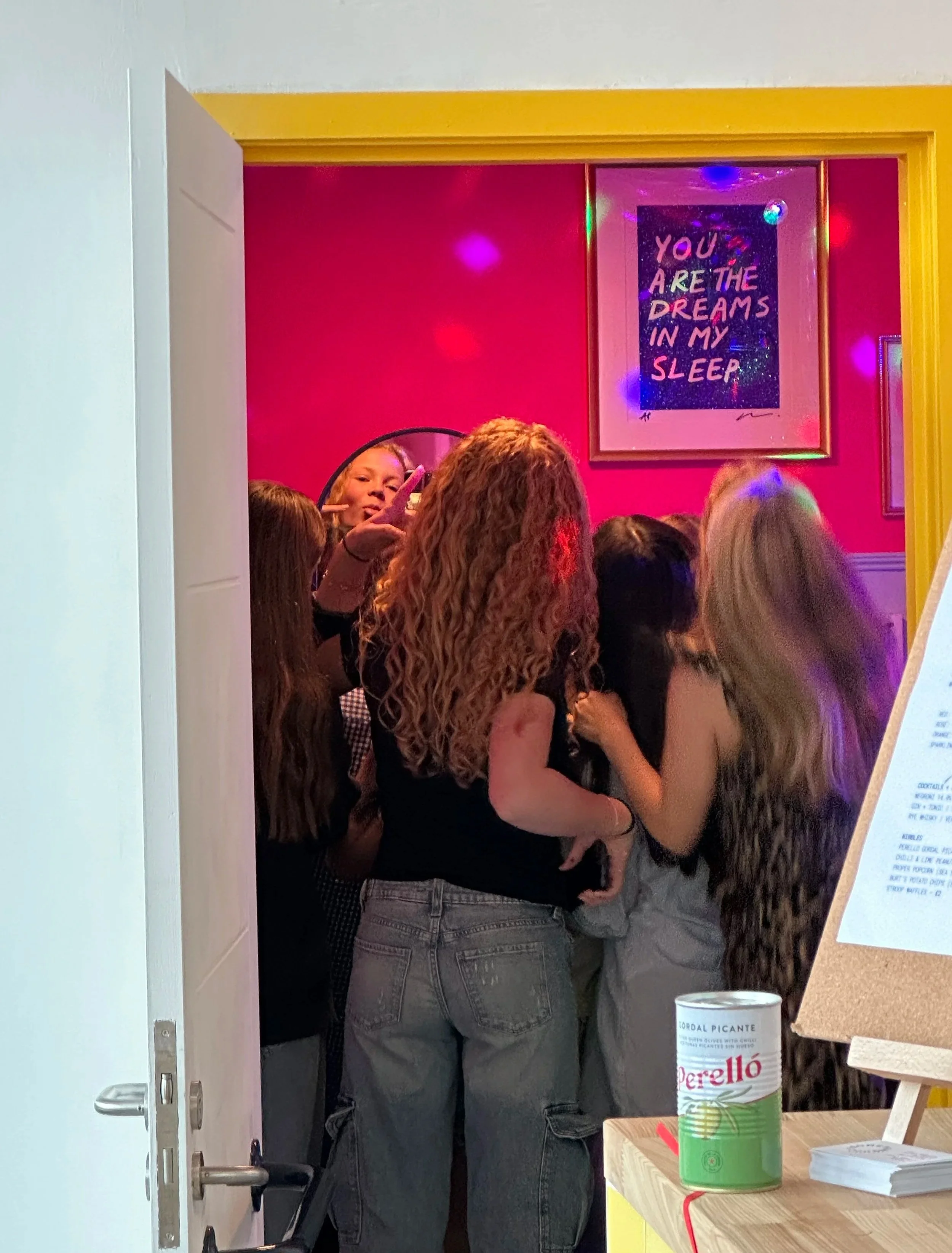 A group of people standing close together inside a room, viewed from the open door. There is a pink wall with a framed sign that says 'You are the dreams in my sleep.' Bright, colorful lights are cast around the room. A woman is visible holding a phone.