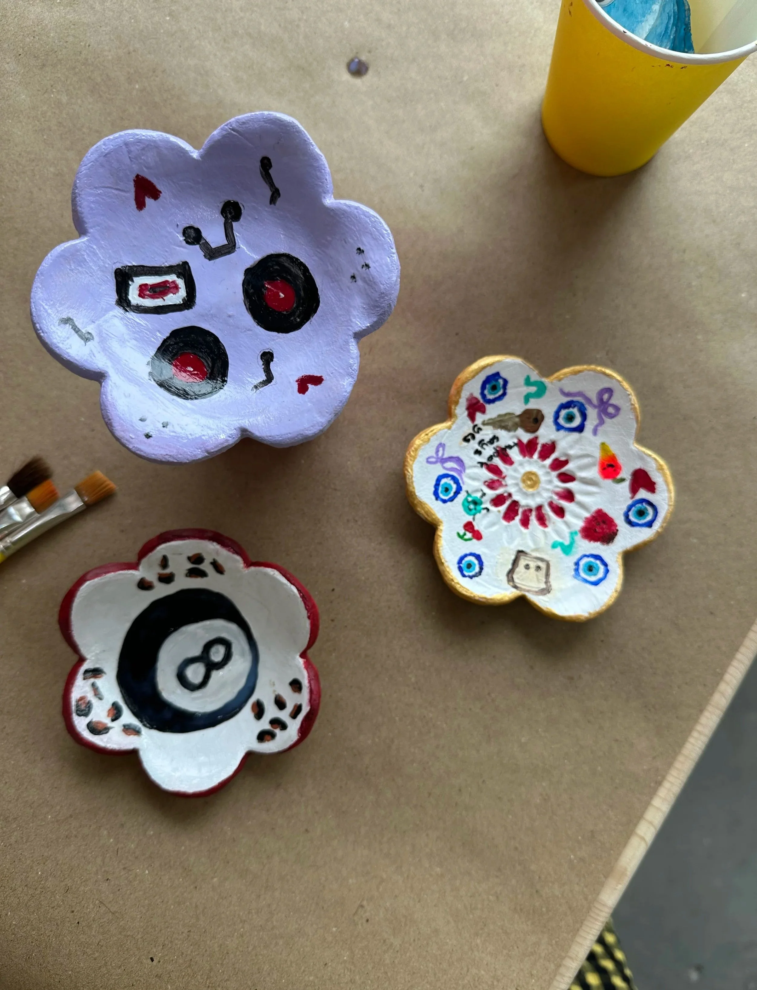 Decorative flower-shaped trinket trays with painted designs, including a smiling face, hearts, and colorful patterns, placed on a beige surface with a yellow cup partially visible in the background at a teen birthday party at Something Good Workshops