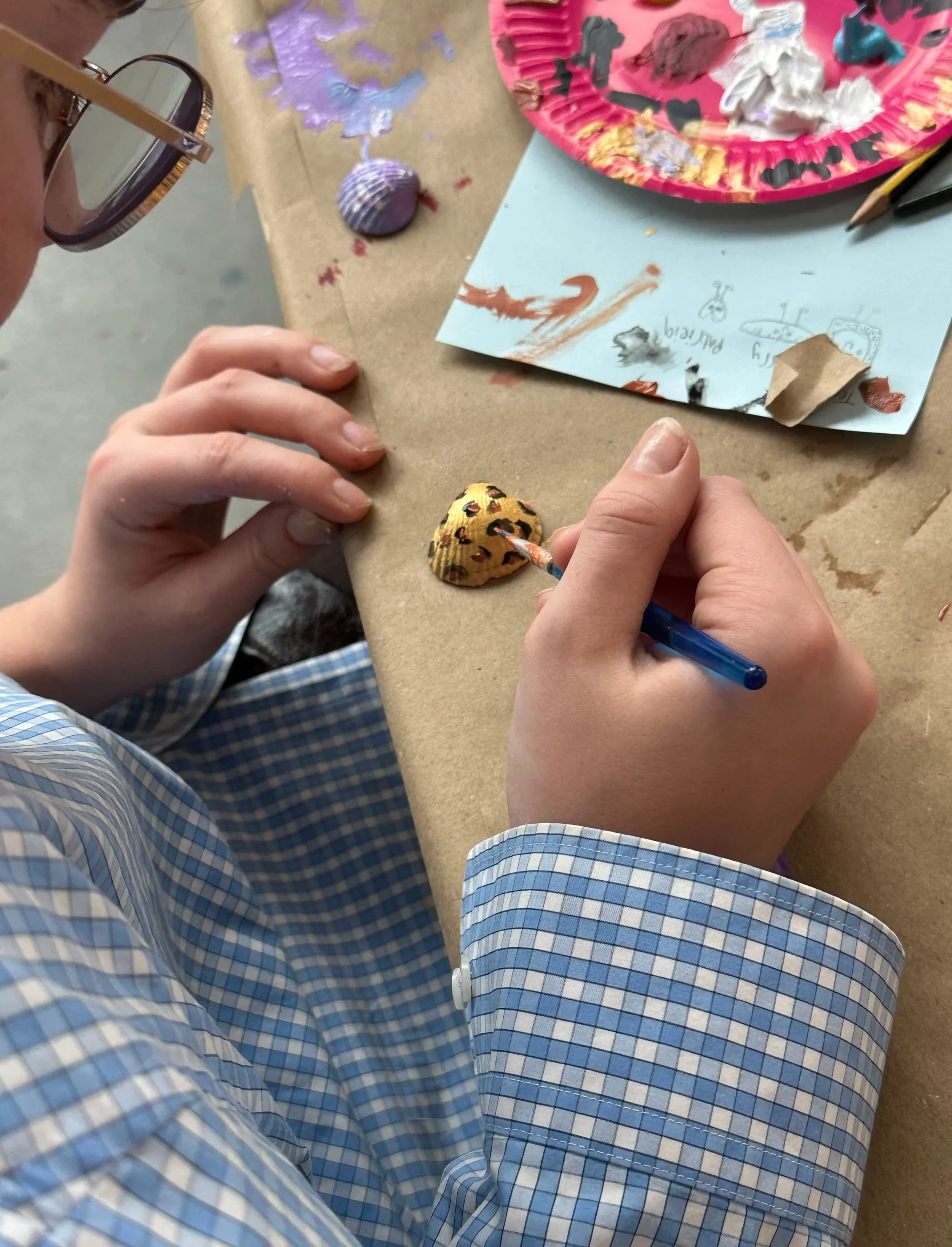 A person's hands painting small shells with black spots and a painted design using a small brush, with a palette of different paint colors nearby. The person is wearing glasses and a blue checkered shirt.