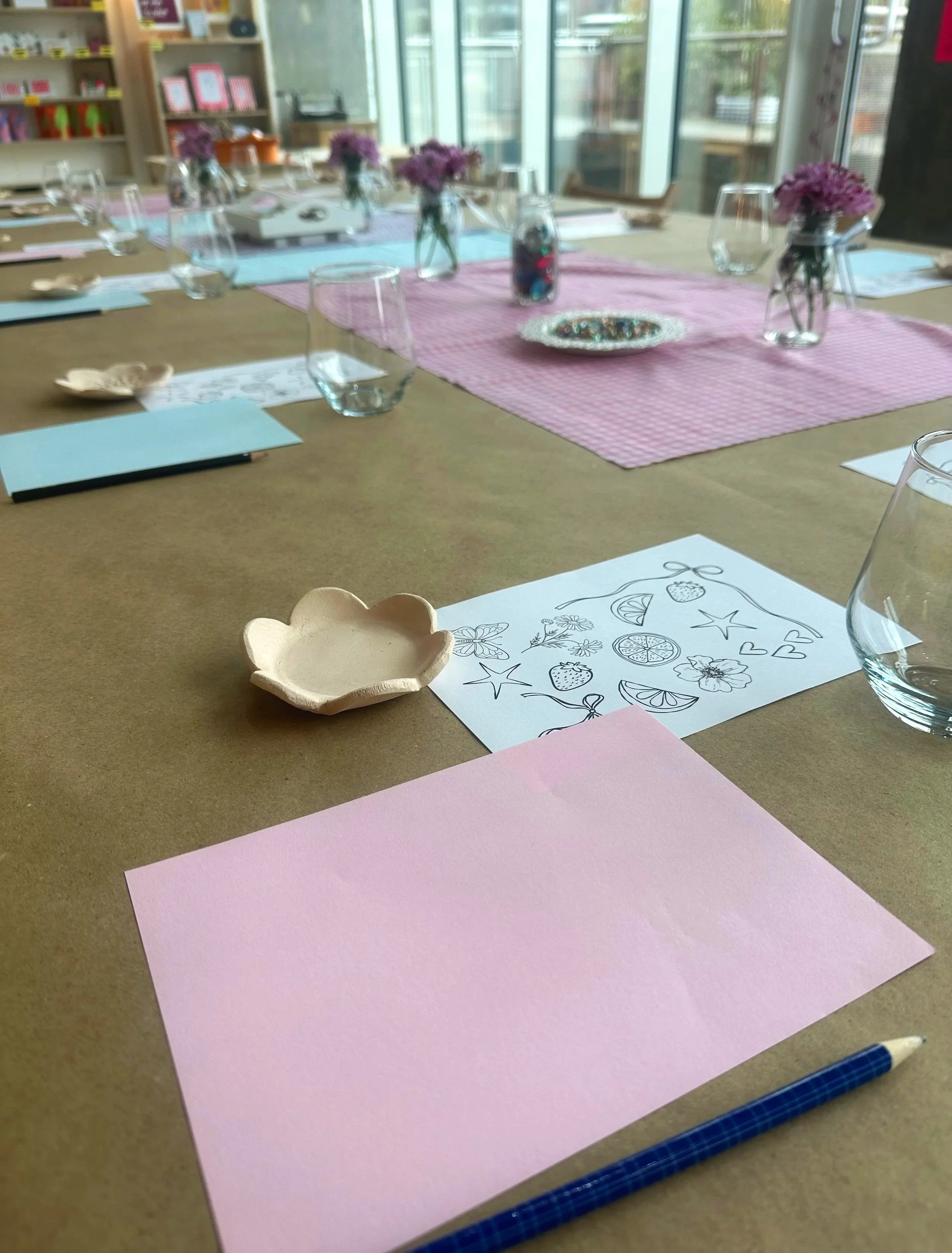 Table set with pink and blue paper, clear glasses, small vases with purple flowers, and a decorative bowl, with a coloring sheet and pen in the foreground.