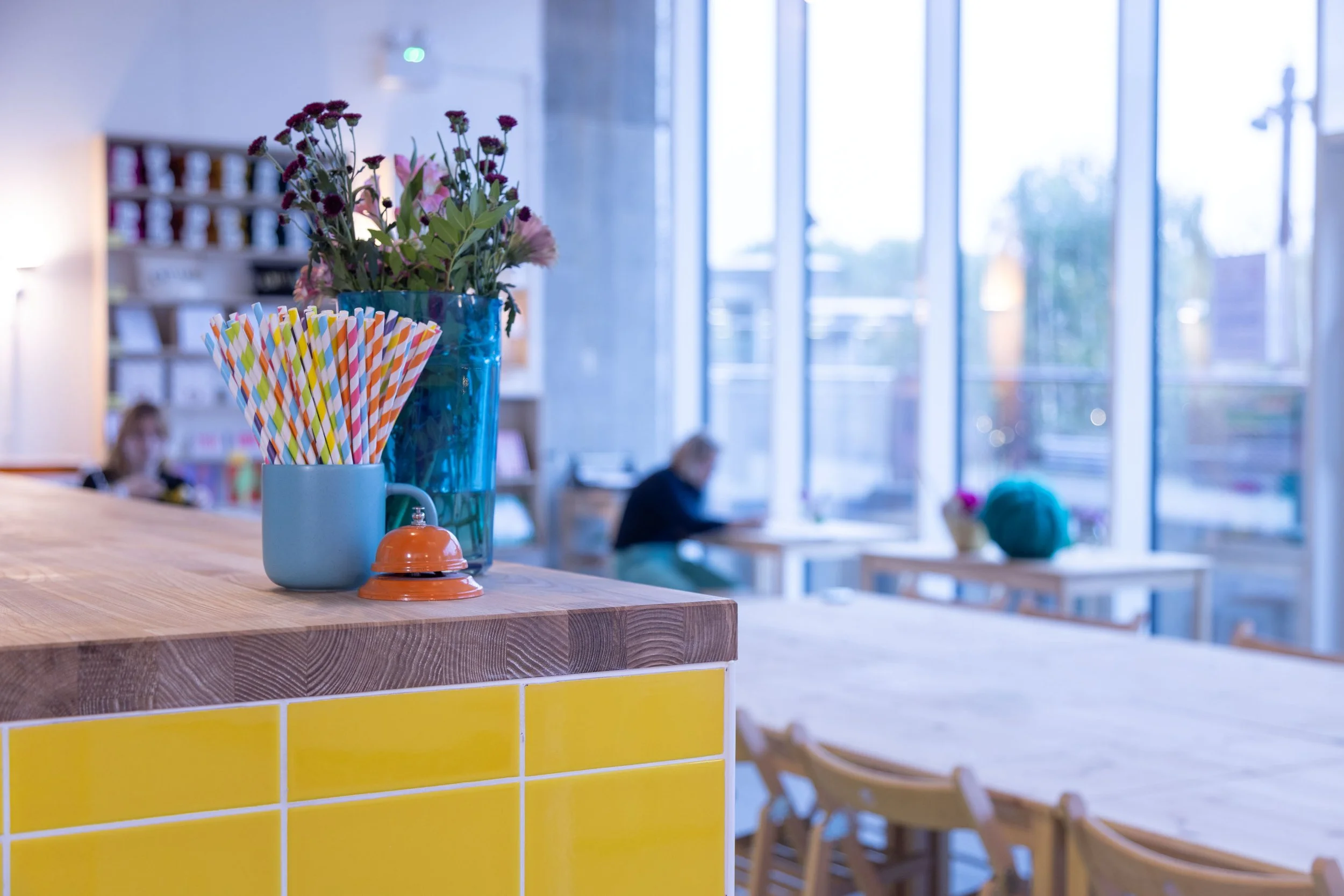 A craft space interior bright with large windows, showing a table with a blue vase of pink and purple flowers and a colorful striped straw holder on a wooden counter with yellow tiles. In the background, blurred customers are seated at wooden tables.