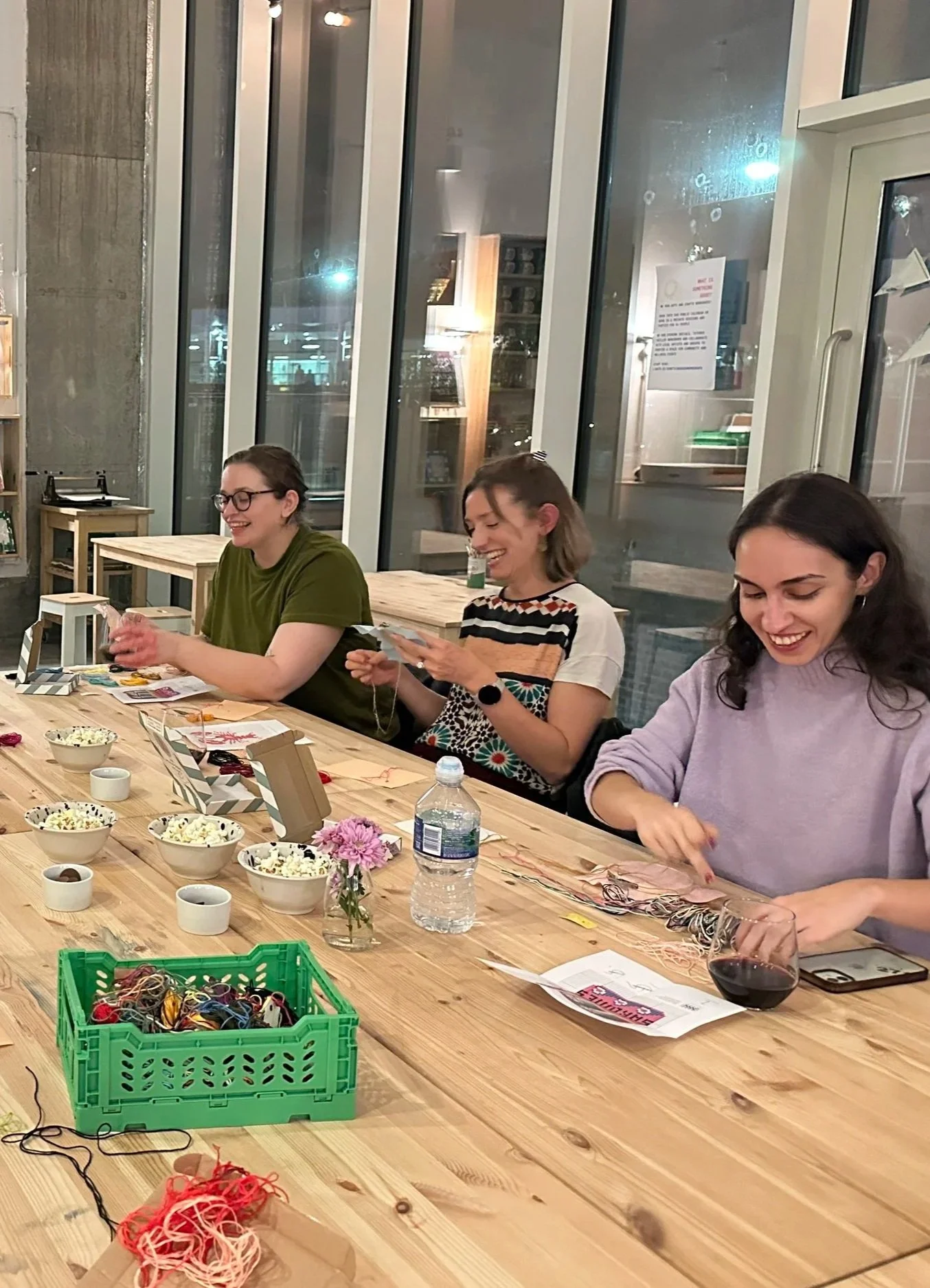 Three women sitting at a wooden table decorated with bowls of popcorn, flowers, and craft supplies, enjoying a social activity in a cozy indoor setting at night.
