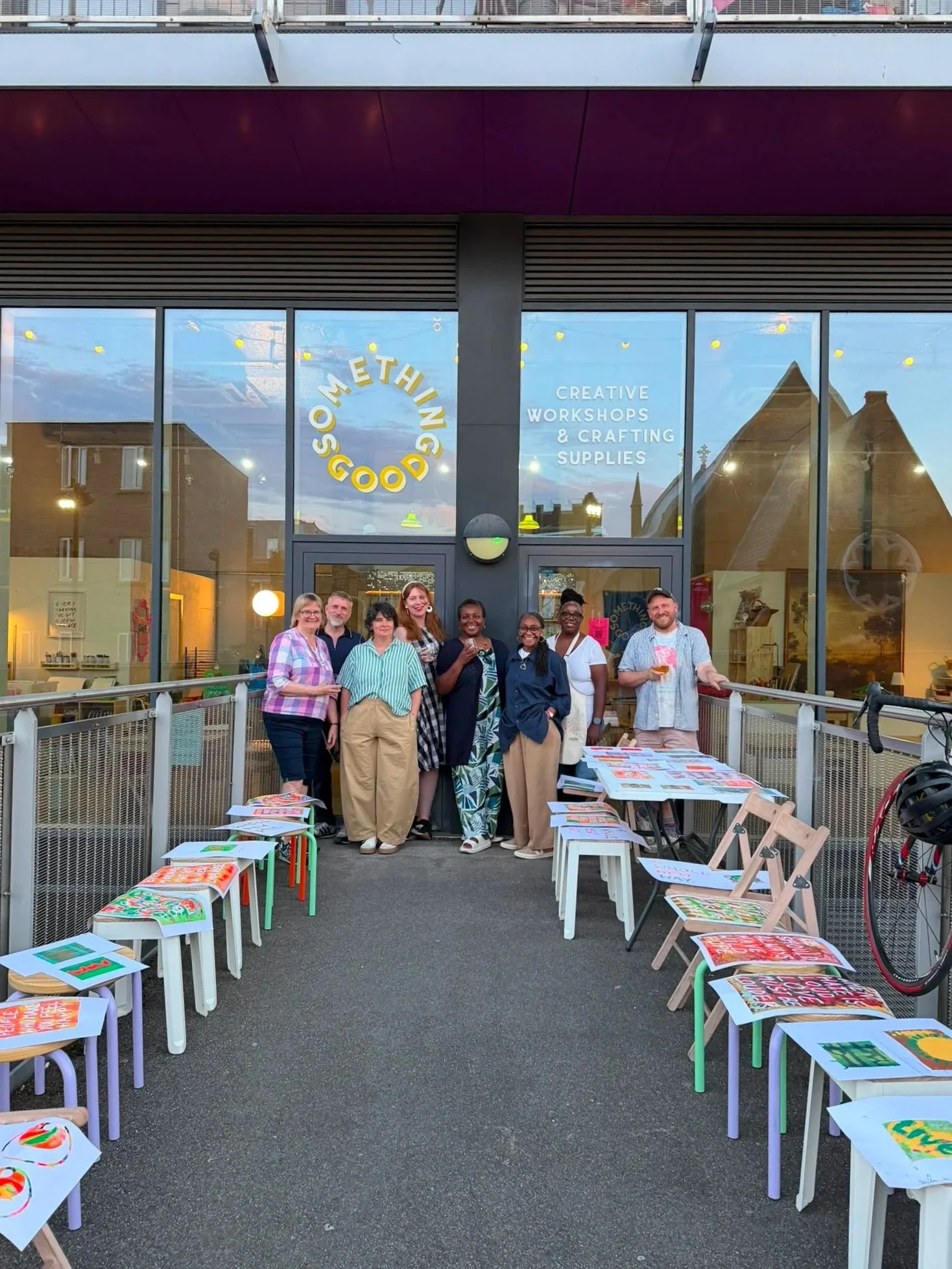 Group of people standing outside a store called 'Some Things Good' with design workshops and crafting supplies, surrounded by chairs and tables with artwork.