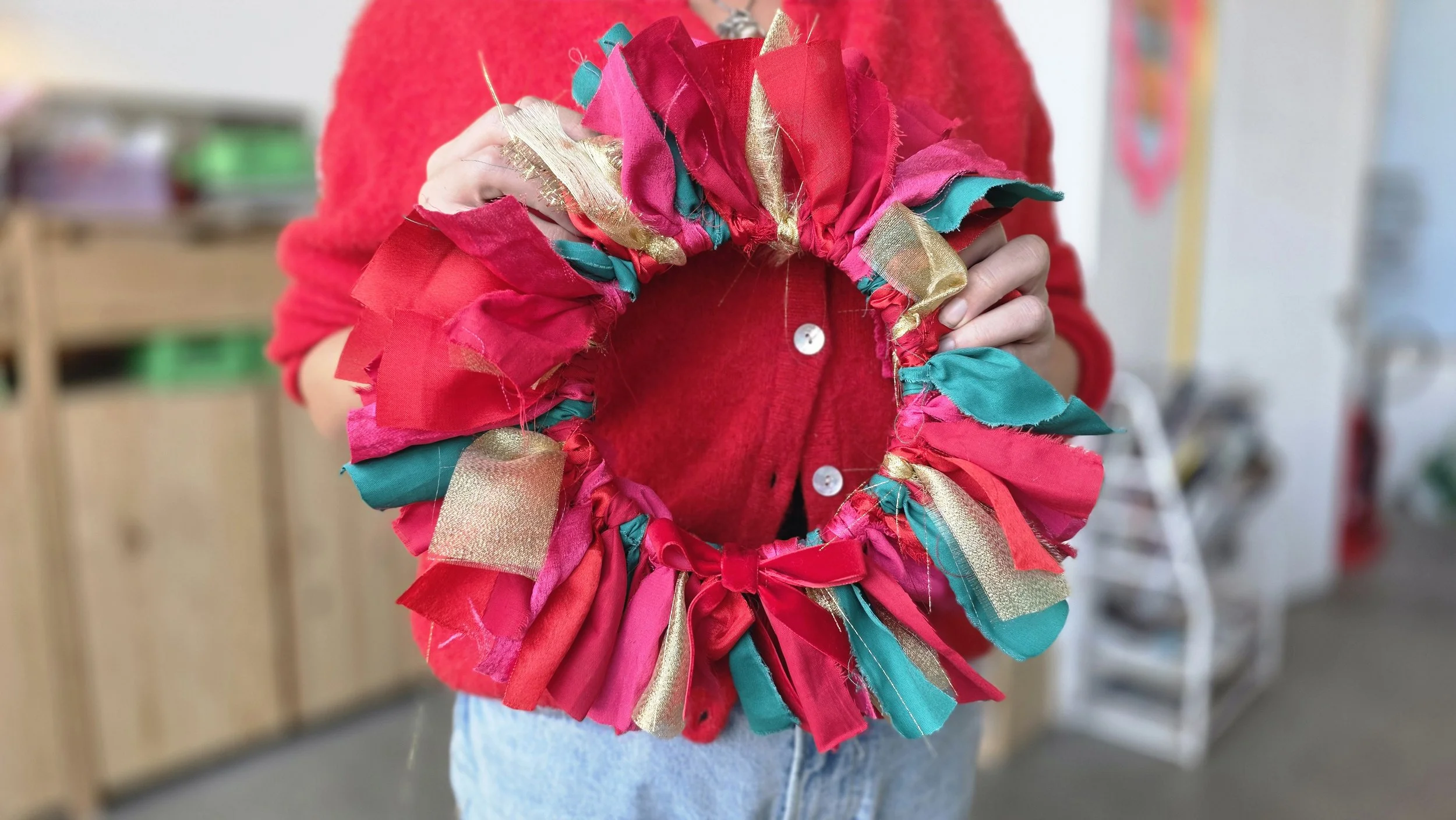Someone in a red jumper holding a colorful holiday wreath made of fabric ribbons in red, teal, and gold.