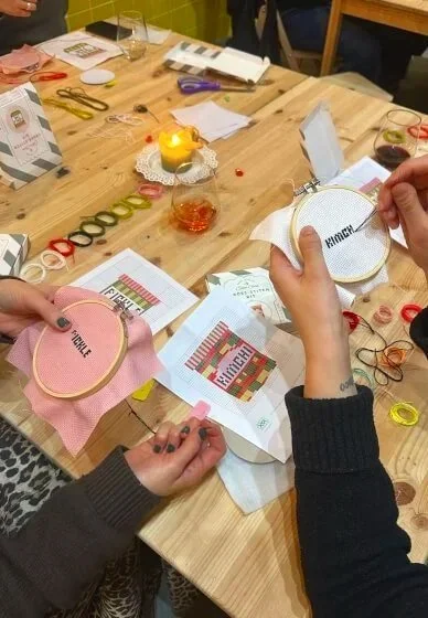People doing cross stitch embroidery at a wooden table with various supplies, including embroidery hoops, colored threads, scissors, and pattern papers.