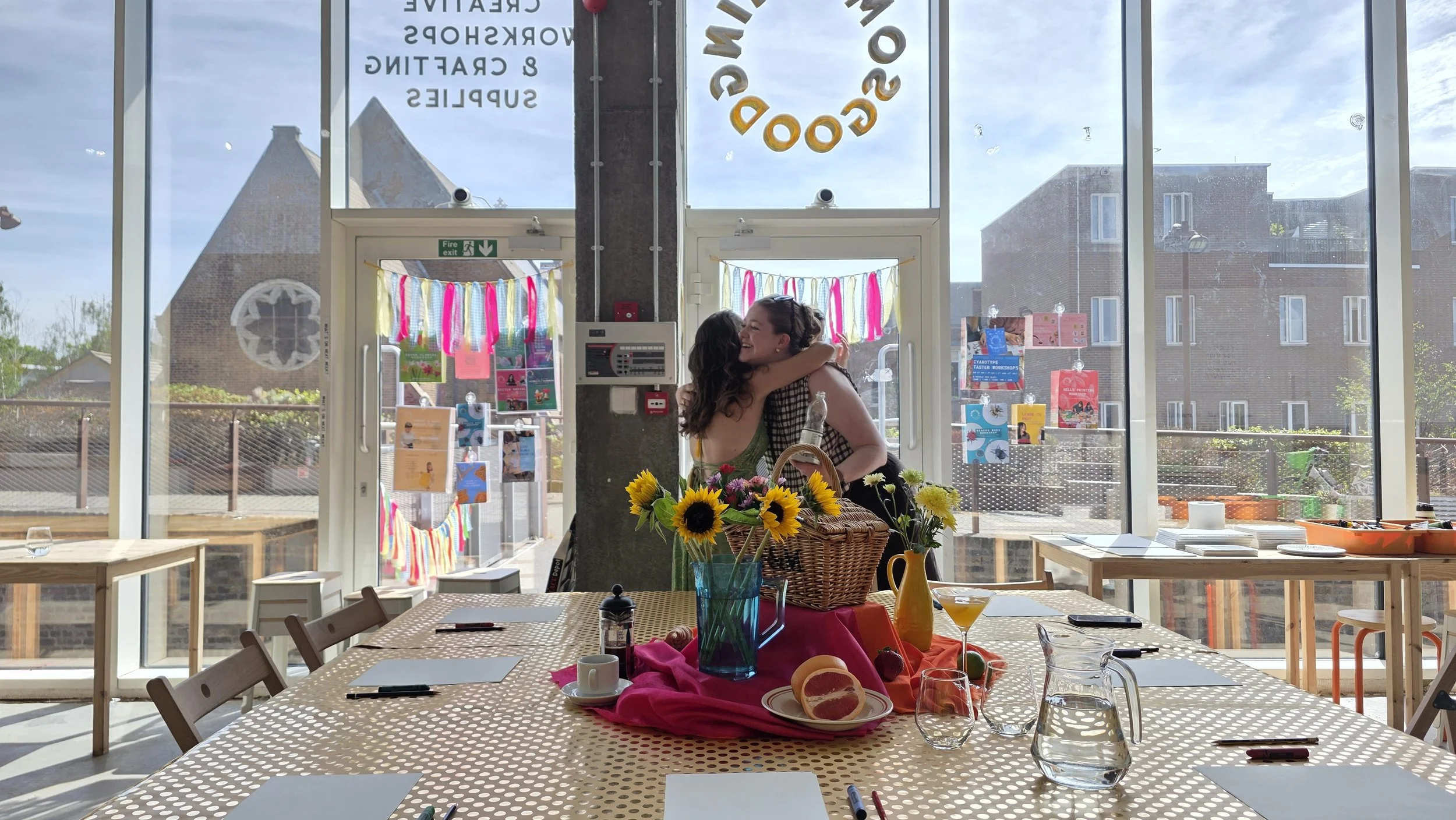 Two women hug inside a bright, decorated craft workshop with large windows showing a church and residential buildings outside. The room has a table with sunflowers, drinks, and various items, indicating a celebration or gathering.