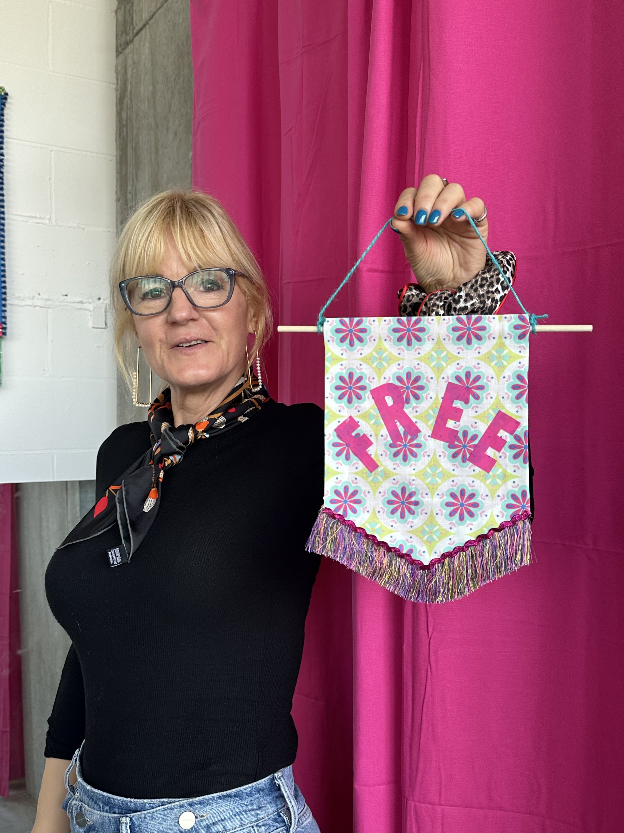 A woman with blonde hair and glasses holding a colorful decorative banner that spells "PEACE" in front of a pink curtain.