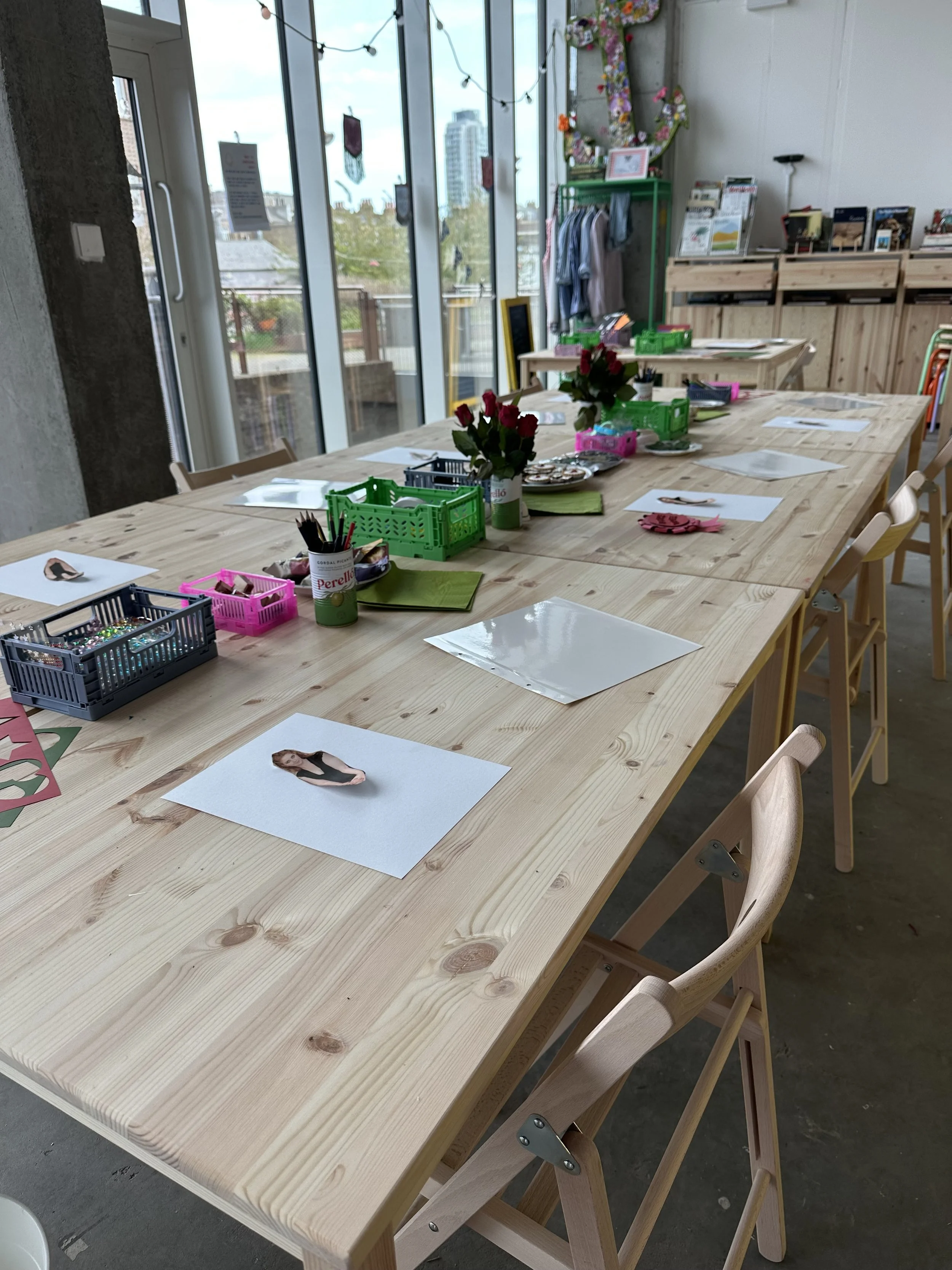 A long wooden table with chairs, decorated with pink roses in vases, various craft supplies, and printed photos, in a bright room with large windows and string lights.