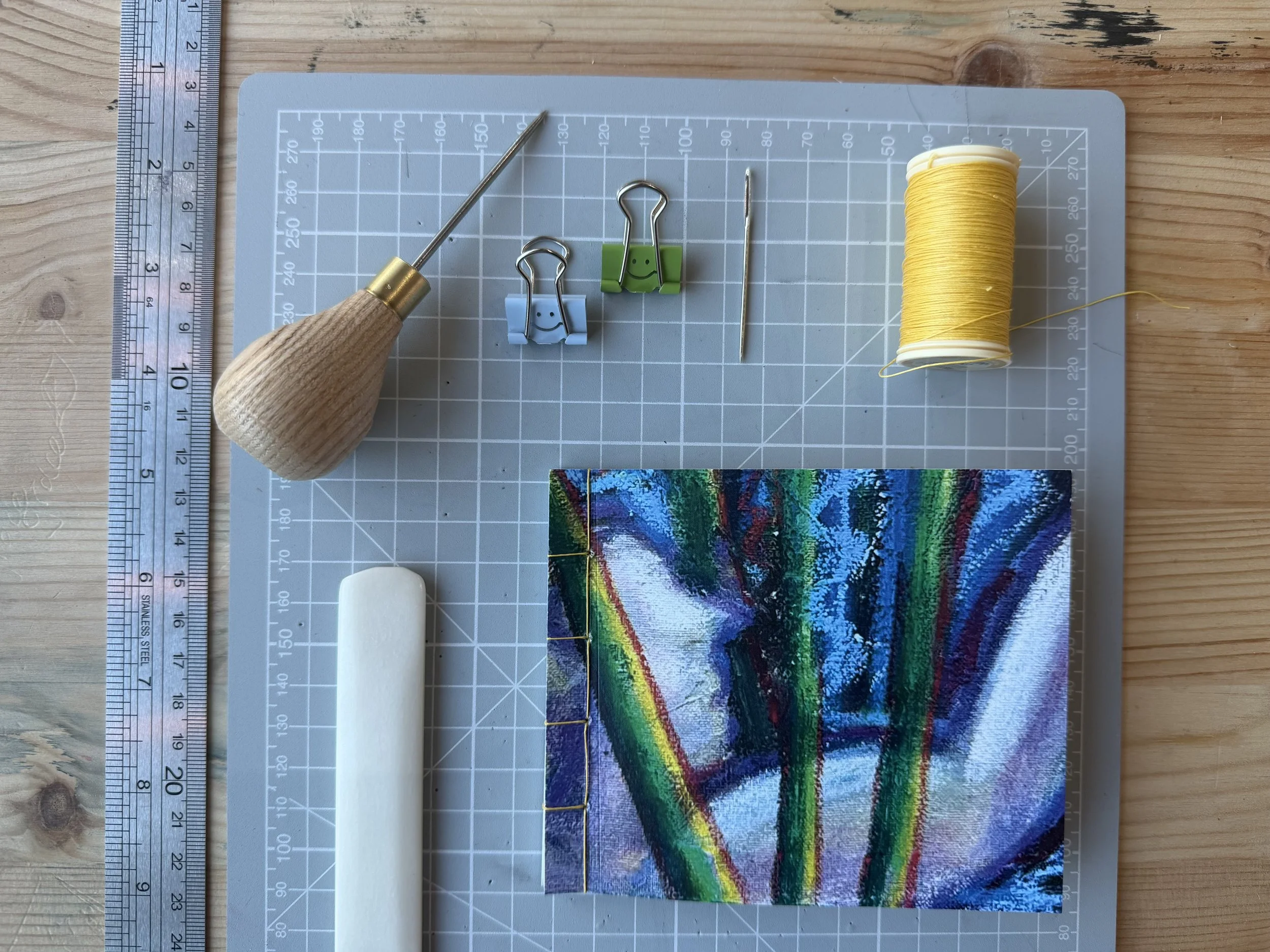 Artwork featuring vibrant rainbow colors, with a wavy pattern on fabric, placed on a cutting mat with measurement markings. Sewing tools and thread are visible around the fabric, on a wooden surface.