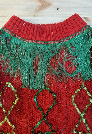 Close-up of a red Christmas sweater with green fringe and bead decorations at a Christmas Jumper Upcycling Workshop in London.