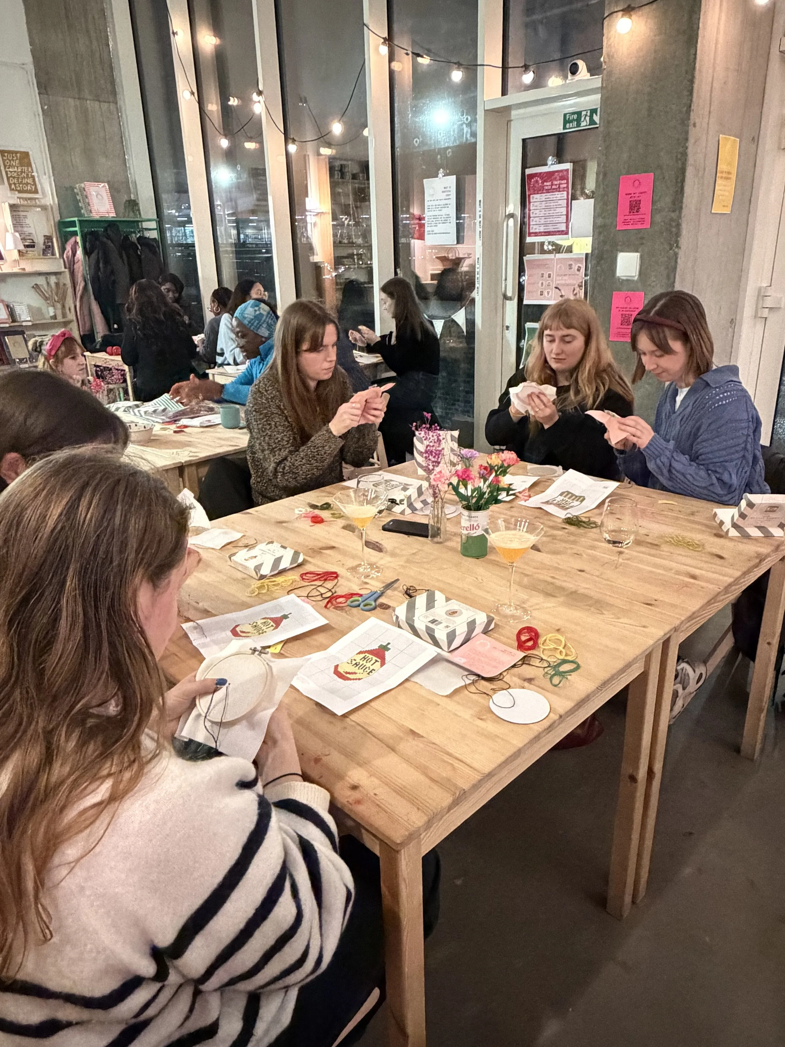 People sitting around a large wooden table engaging in crafts or sewing activities inside a cozy, well-lit room with large windows, string lights, and various decorations.