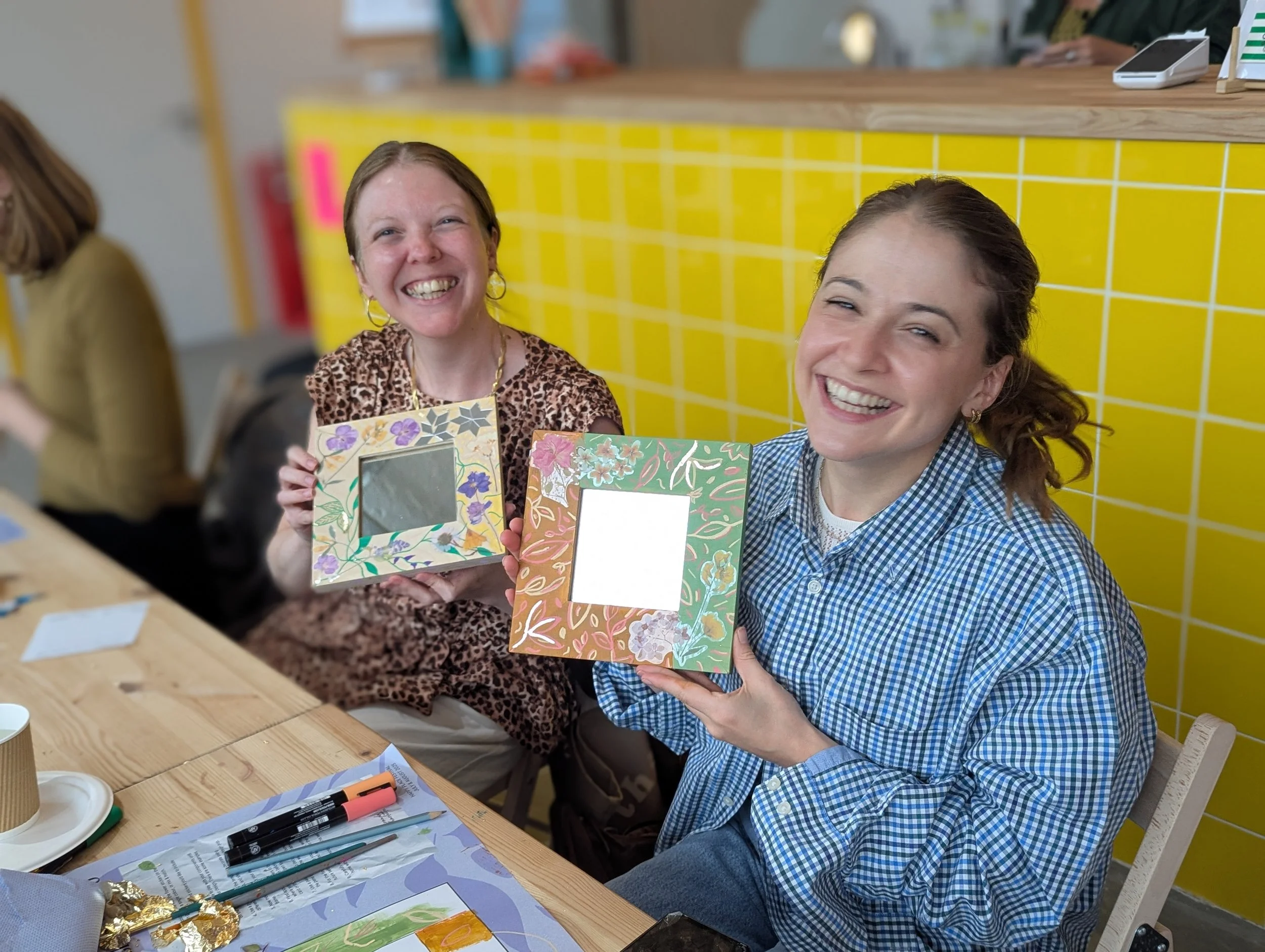 Two women holding decorated photo frames and smiling, sitting at a wooden table with art supplies in a colorful, casual setting.